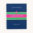 The image shows A Simplified Life by Emily Ley, from the brand Simplified. The dark blue cover features pink and green stripes, with the subtitle tactical tools for intentional living, offering tips for streamlining routines.