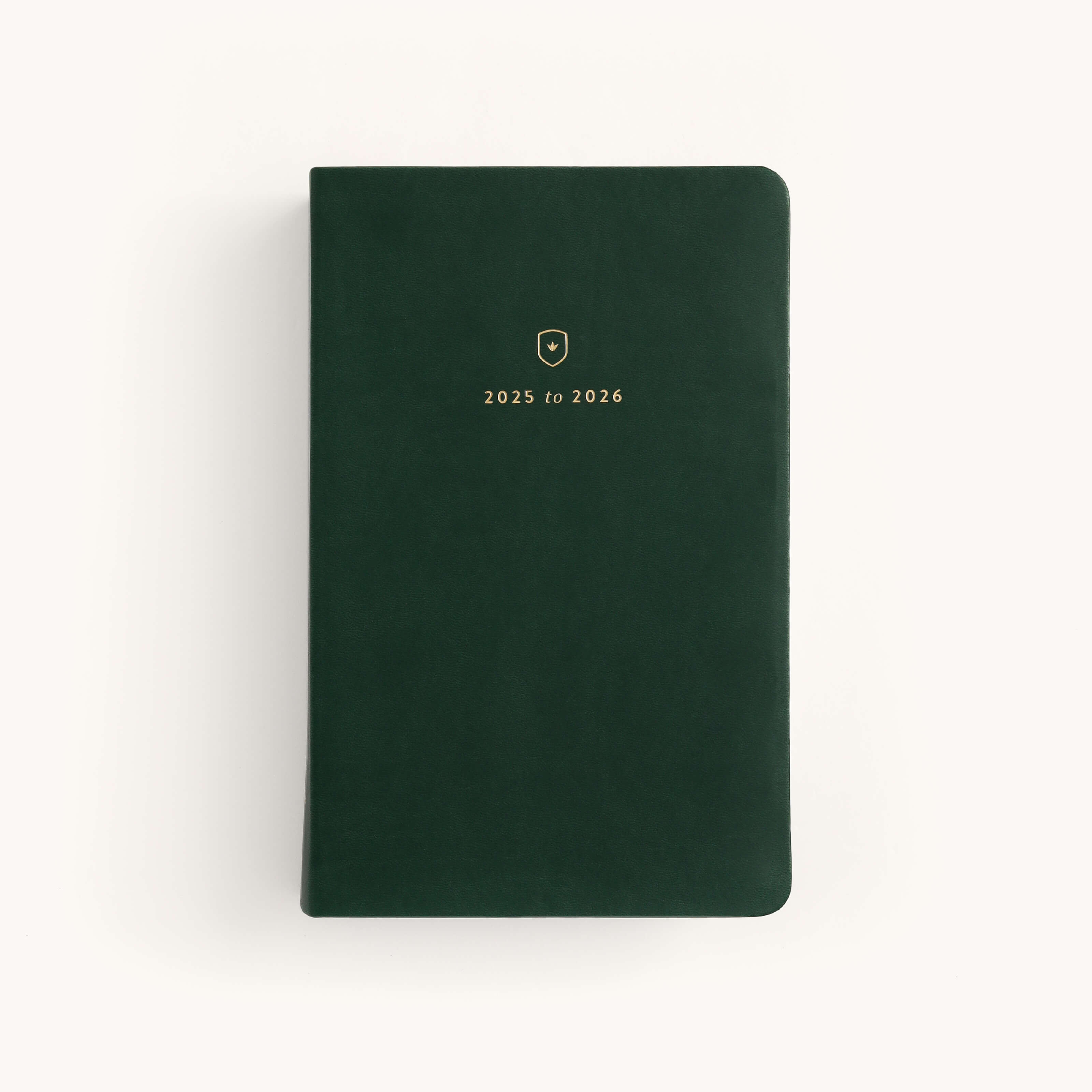 The Simplified 2025-2026 Bookbound Daily Dapperdesk Planner in Evergreen features a green leather-bound cover with rounded corners. It includes a small gold emblem and 2025 to 2026 text in gold, centered on the front, making it perfect for detailed planning against a light background.