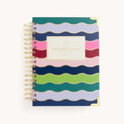 A vibrant spiral-bound planner with wavy stripes in blue, green, pink, and purple enhanced by gold foil. The central label reads 2025-2026 Spiral Daily Simplified Planner - Rainbow Row, ideal for organizing your Simplified Prep Work.