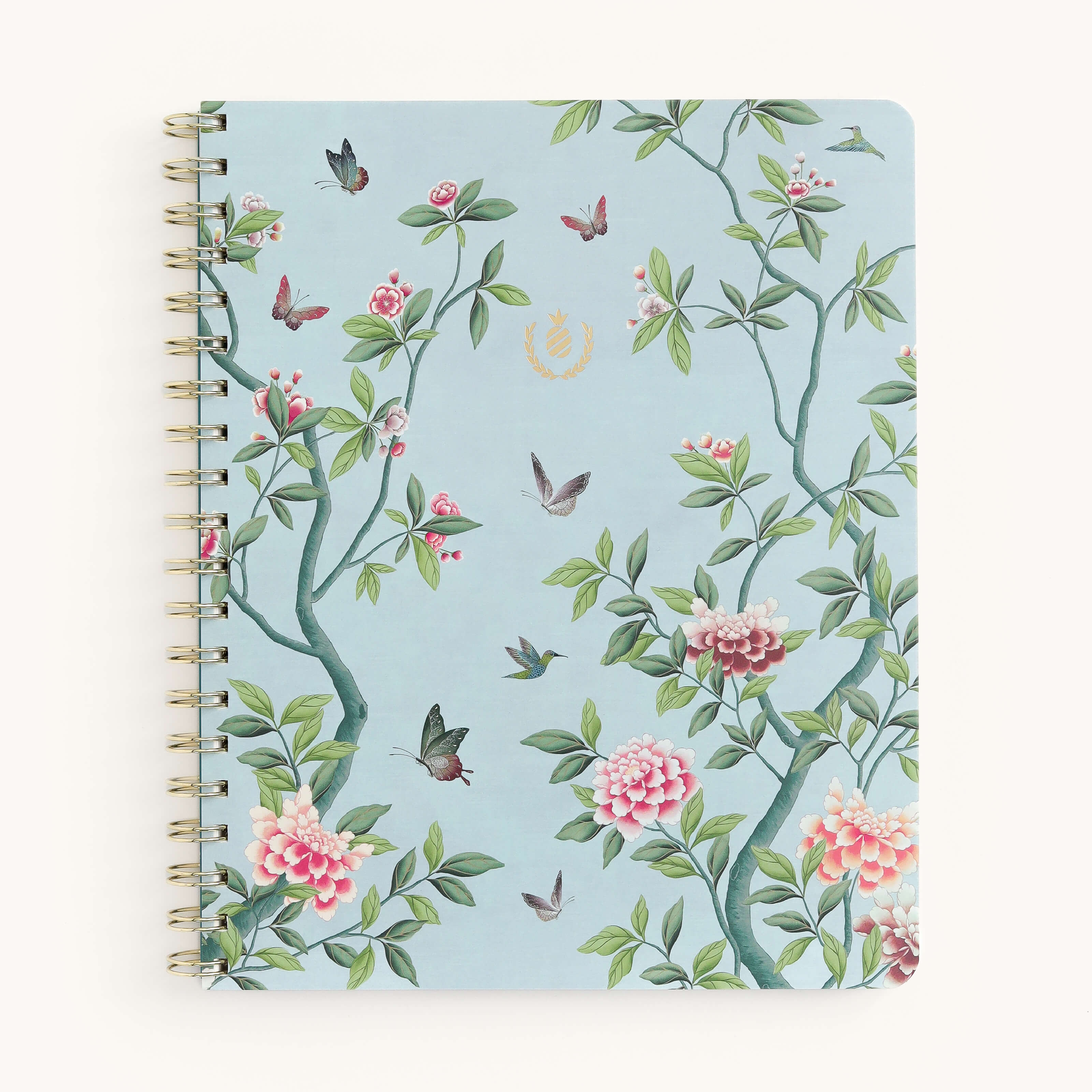 The Notebook - Faraway Land by Simplified features a soft-touch light blue cover with pink and white floral patterns, green leaves, and flying birds. A gold wire-o binding complements the elegant gold emblem centered near the top.