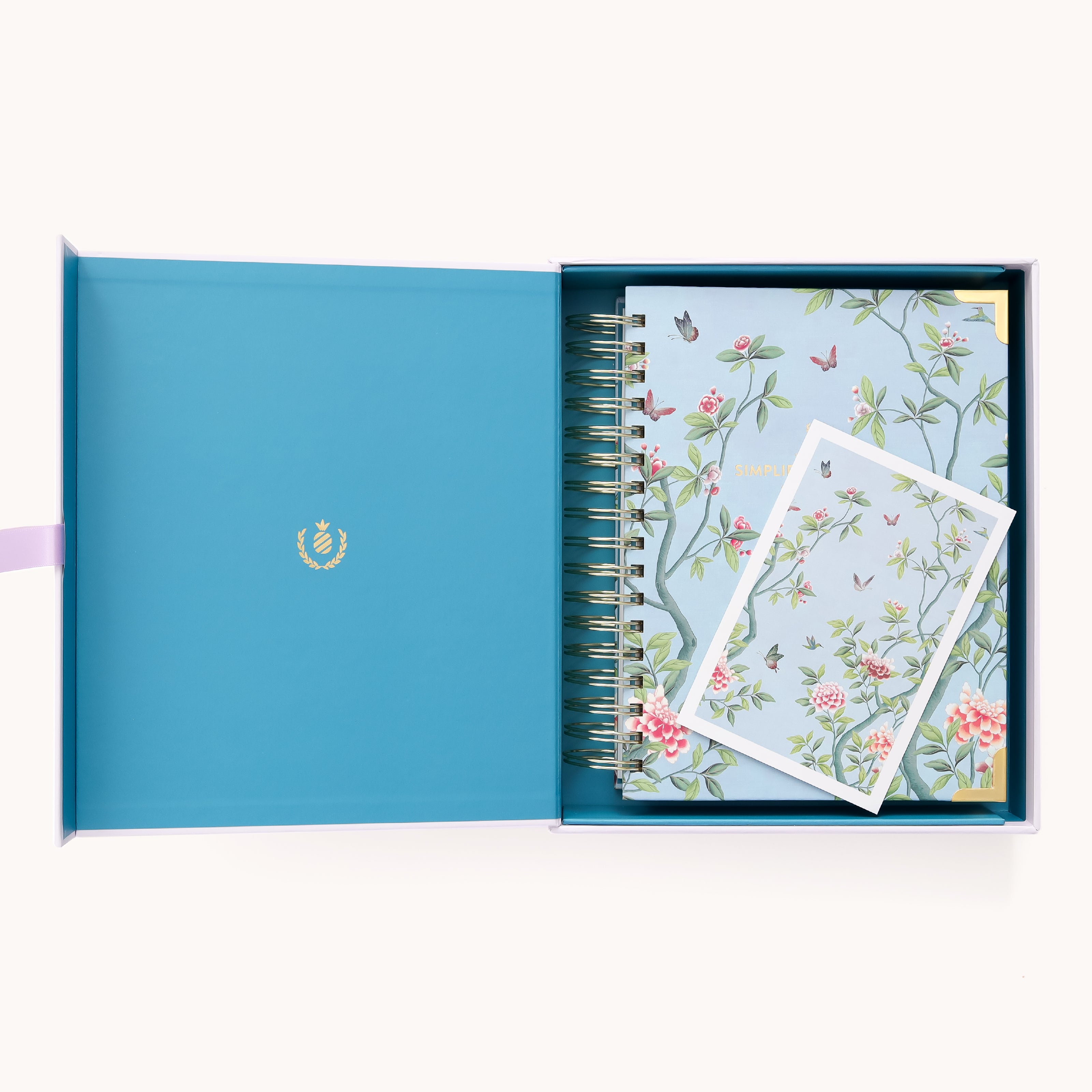The open blue gift box holds the 2026 Spiral Daily Simplified Planner - Faraway Land and a matching card by Simplified, both with hand-painted botanical and bird designs on a light blue background. The lid’s inside has a small gold emblem.