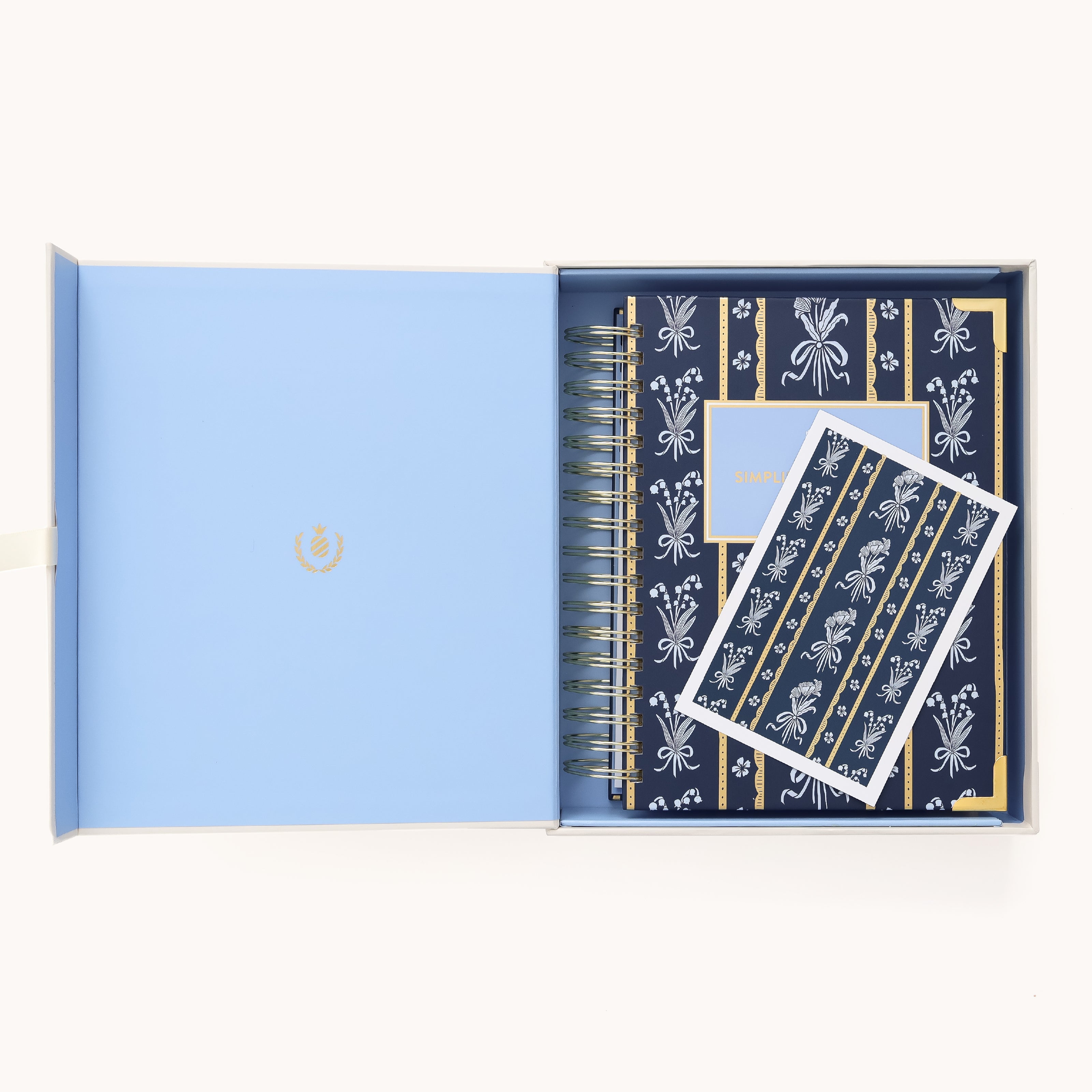 An open blue box shows the 2026 Spiral Daily Simplified Planner - Poppy Posies by Simplified and a matching card, both with navy covers, white bows, and gold accents in a striped design—ideal for your 2026 planning needs.