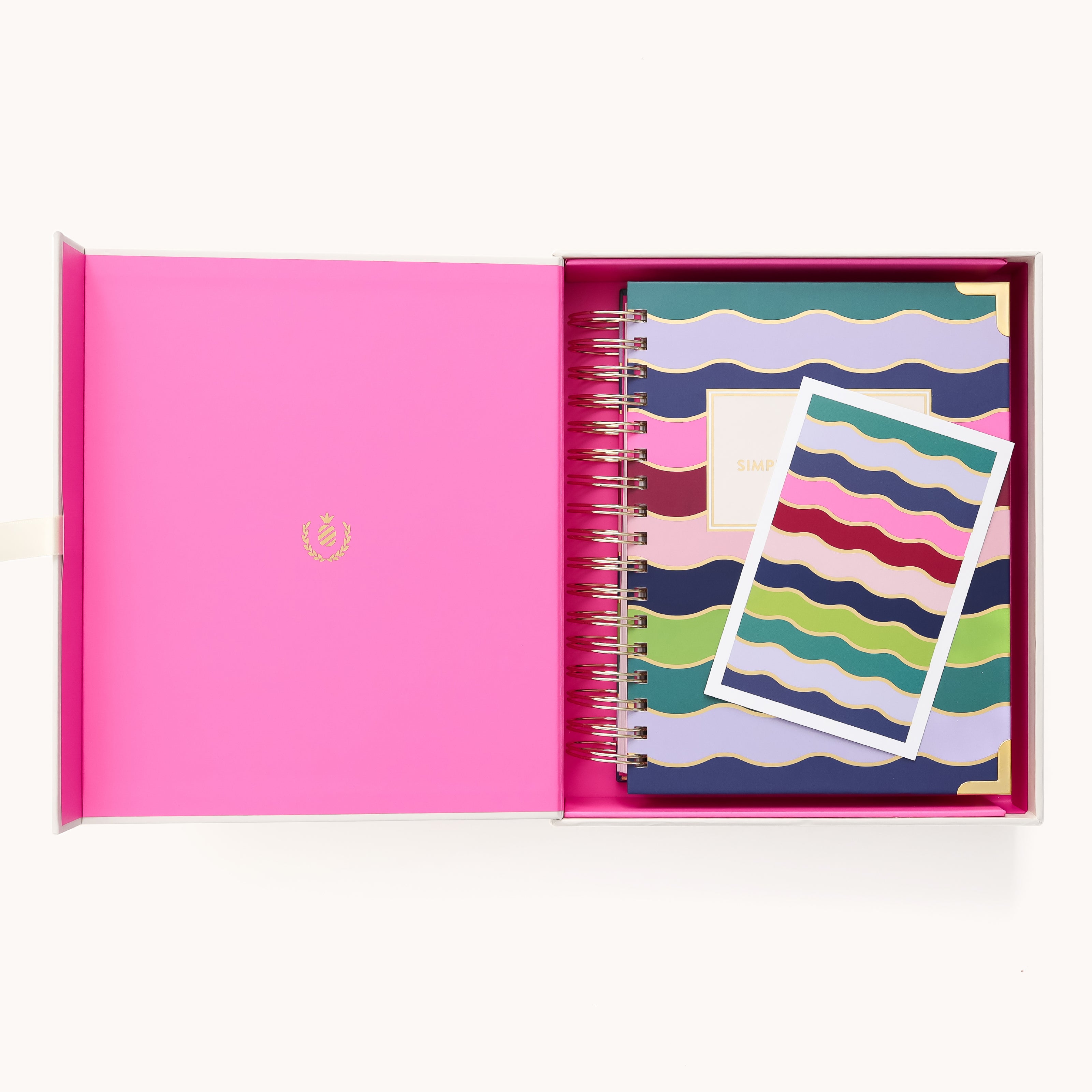 An open pink box showcases the Simplified 2026 Spiral Daily Simplified Planner – Rainbow Row and a coordinating postcard, both with vibrant wavy patterns. The box lid features a gold emblem. Perfect for daily organization in signature Simplified style.