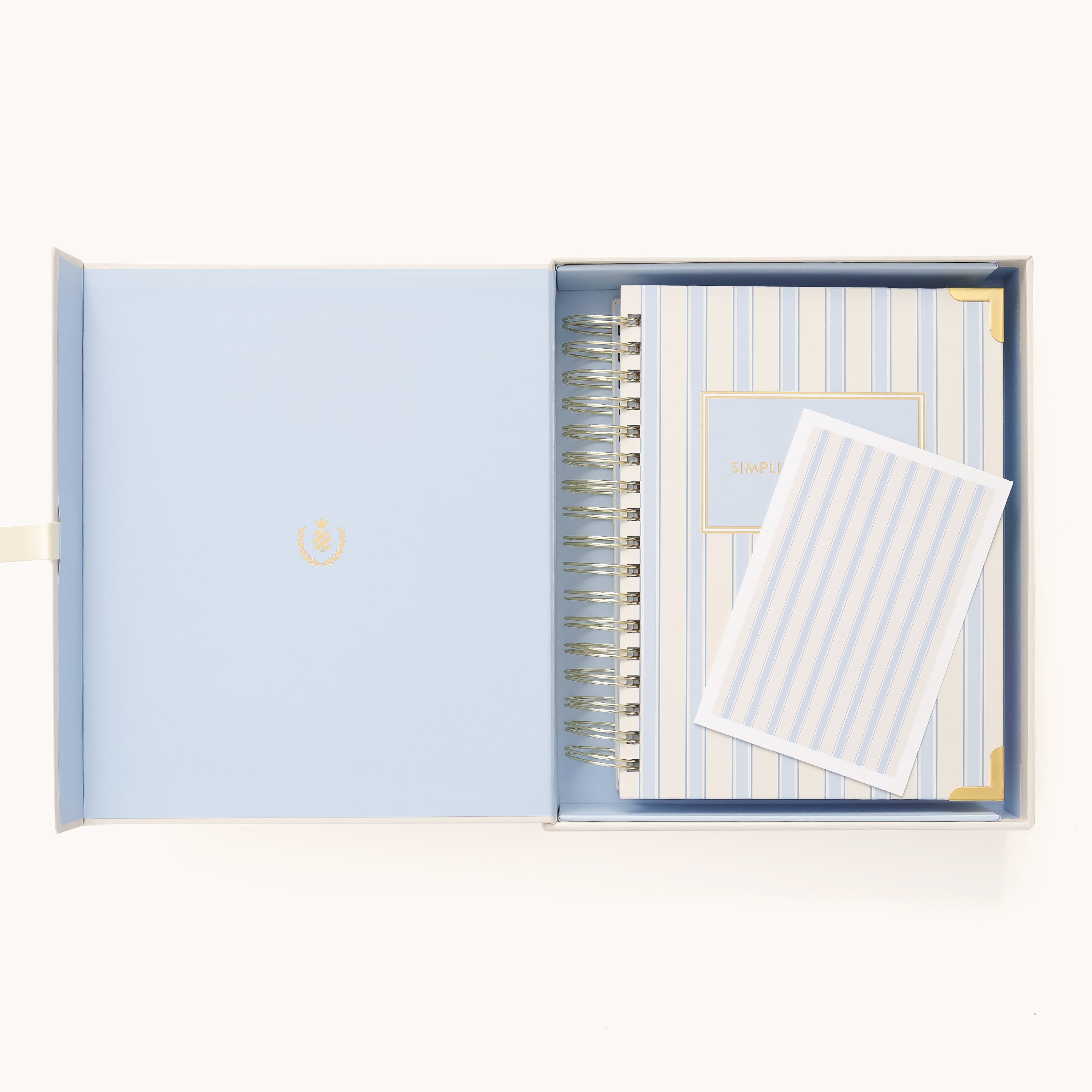 An open light blue box holds a Simplified 2026 Spiral Daily Simplified Planner in Serene Stripe and a matching notepad, both with white and blue vertical stripes and gold accents—the perfect coordinated set.