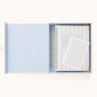 An open light blue box holds a Simplified 2026 Spiral Daily Simplified Planner in Serene Stripe and a matching notepad, both with white and blue vertical stripes and gold accents—the perfect coordinated set.