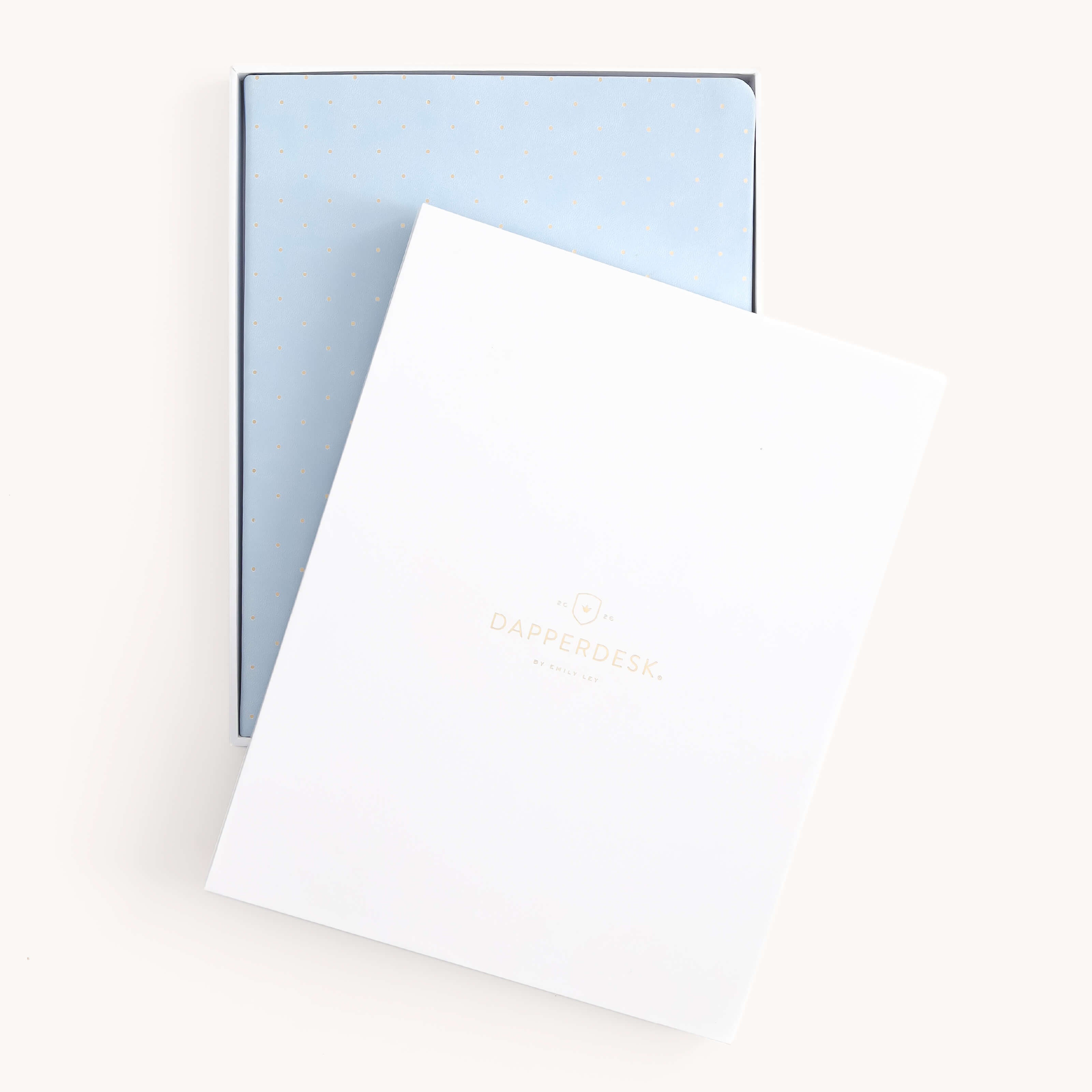The 2026 Bookbound Weekly Dapperdesk Planner - Carolina Dot by Simplified, featuring a light blue leatherette cover with gold dots, is partially covered by a white folder with Dapperdesk in gold on the front.