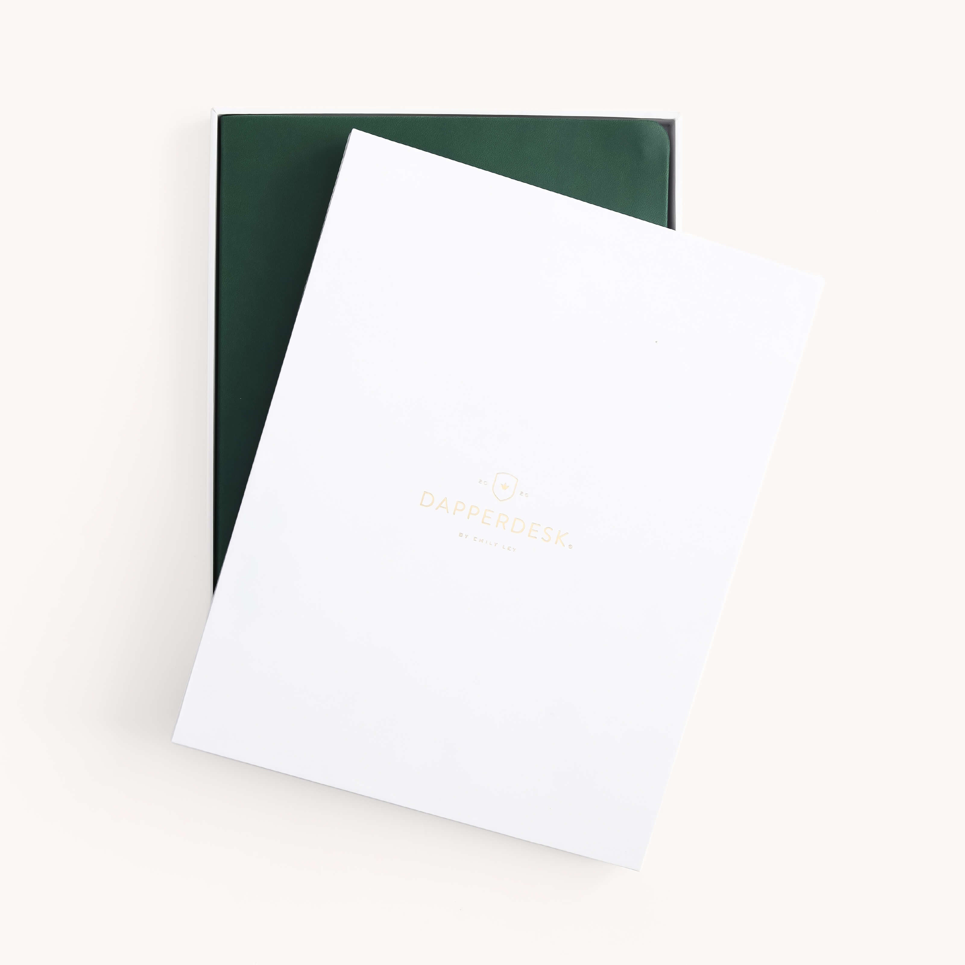 A white folder with DAPPERDESK in gold lettering partially covers the Simplified 2026 Bookbound Weekly Dapperdesk Planner in Evergreen, featuring a leatherette cover, displayed inside an open box on a plain light background.