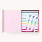 Open box with a pink interior holds the 2025-2026 Bookbound Weekly Simplified Planner - Happy Stripe by Simplified, featuring colorful horizontal stripes and a gold-bordered label. On top is a matching striped card with elegant gold foil details.