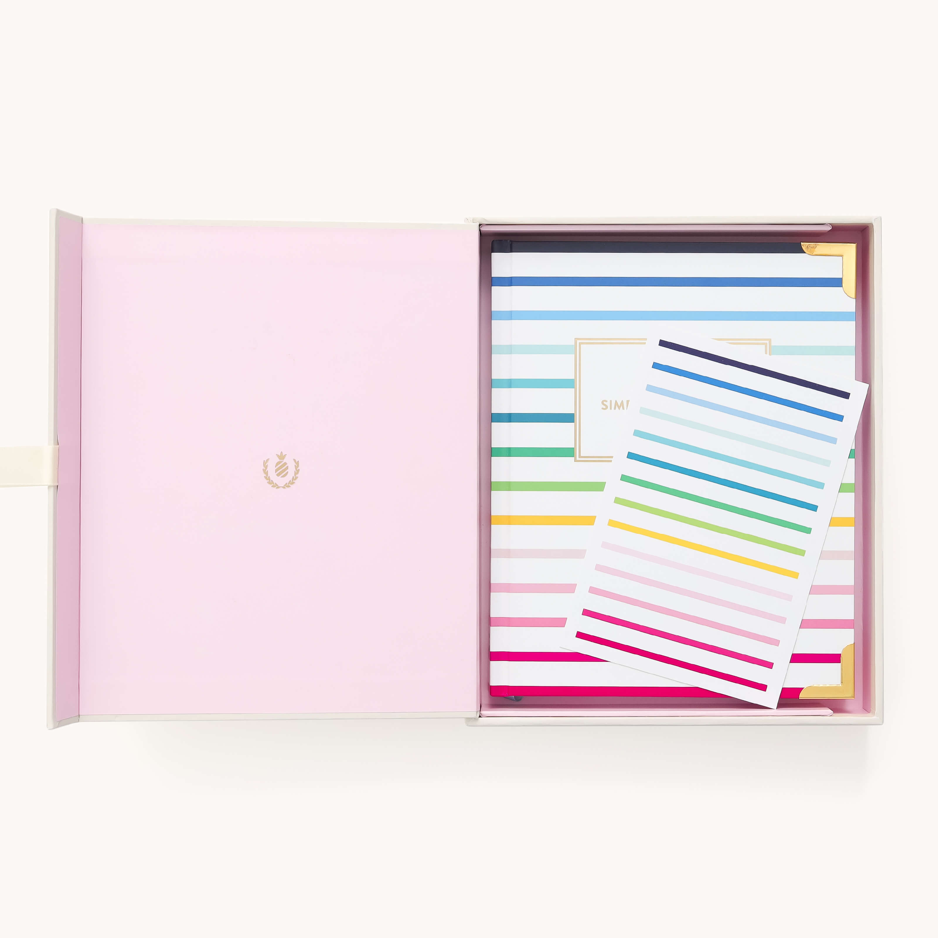 Open box with a pink interior holds the 2025-2026 Bookbound Weekly Simplified Planner - Happy Stripe by Simplified, featuring colorful horizontal stripes and a gold-bordered label. On top is a matching striped card with elegant gold foil details.