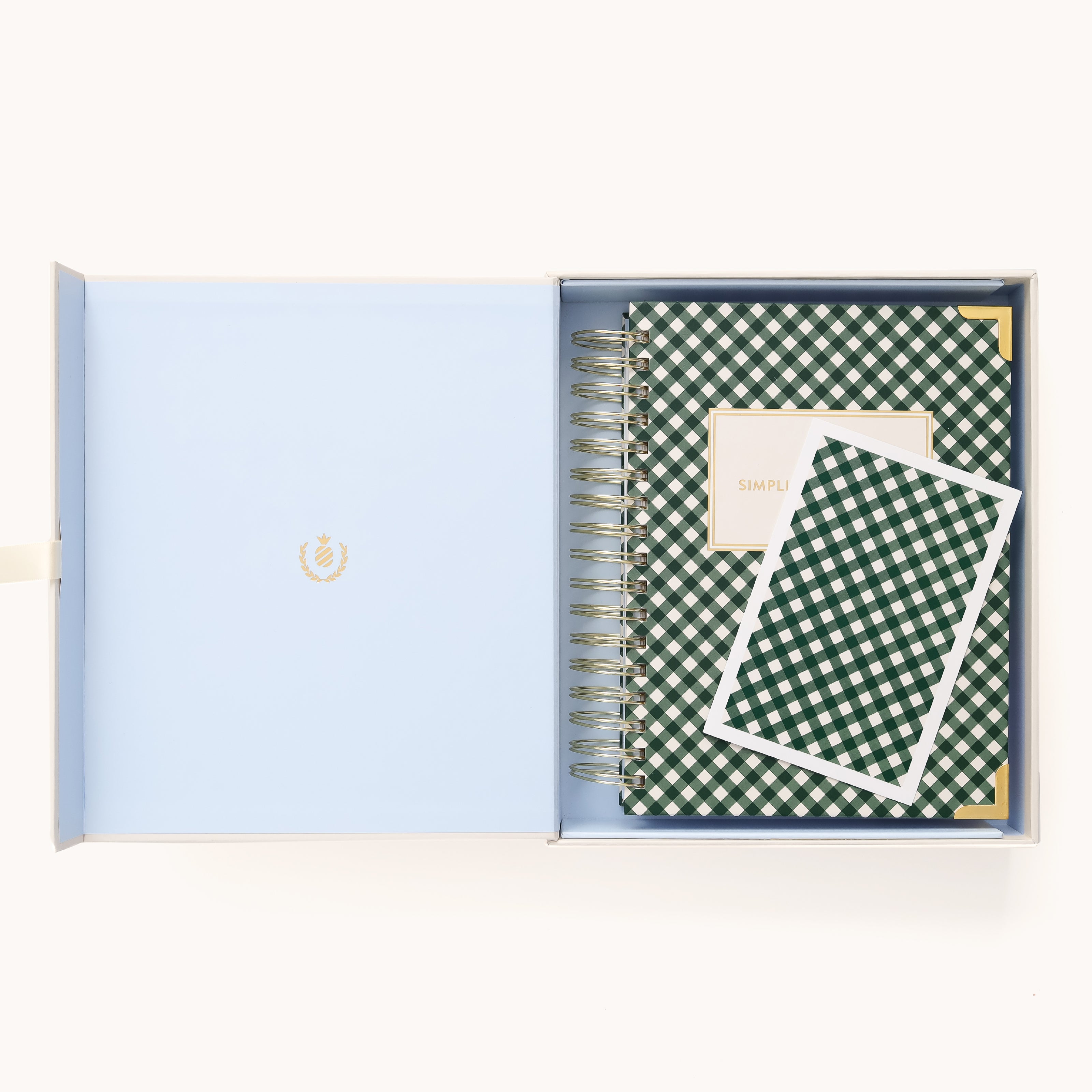 The opened box displays a blue interior with the 2025-2026 Spiral Daily Simplified Planner - Camp Gingham and a small card, both in a green and white checkered pattern. The Simplified Planner features square labels, while gold foil details adorn the emblem on the box lid.