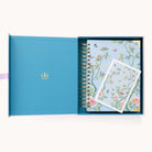 An open box showcases the 2025-2026 Spiral Daily Simplified Planner – Faraway Land by Simplified, featuring a floral and bird design with pink flowers and birds on a light blue background, highlighted by gold foil. A matching card accompanies it, and a gold emblem decorates the lids interior.