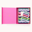 A pink-interior open box reveals the 2025-2026 Spiral Daily Simplified Planner - Rainbow Row by Simplified, with wavy, colorful patterns and a matching card. The planners emblem boasts gold foil details for an elegant touch aimed at fans of streamlined preparations.