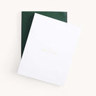 A partially open white box featuring Dapperdesk in gold on the lid reveals a dark green interior, designed to house the 2025-2026 Simplified Bookbound Weekly Dapperdesk Planner - Evergreen, resting elegantly against a light neutral backdrop.
