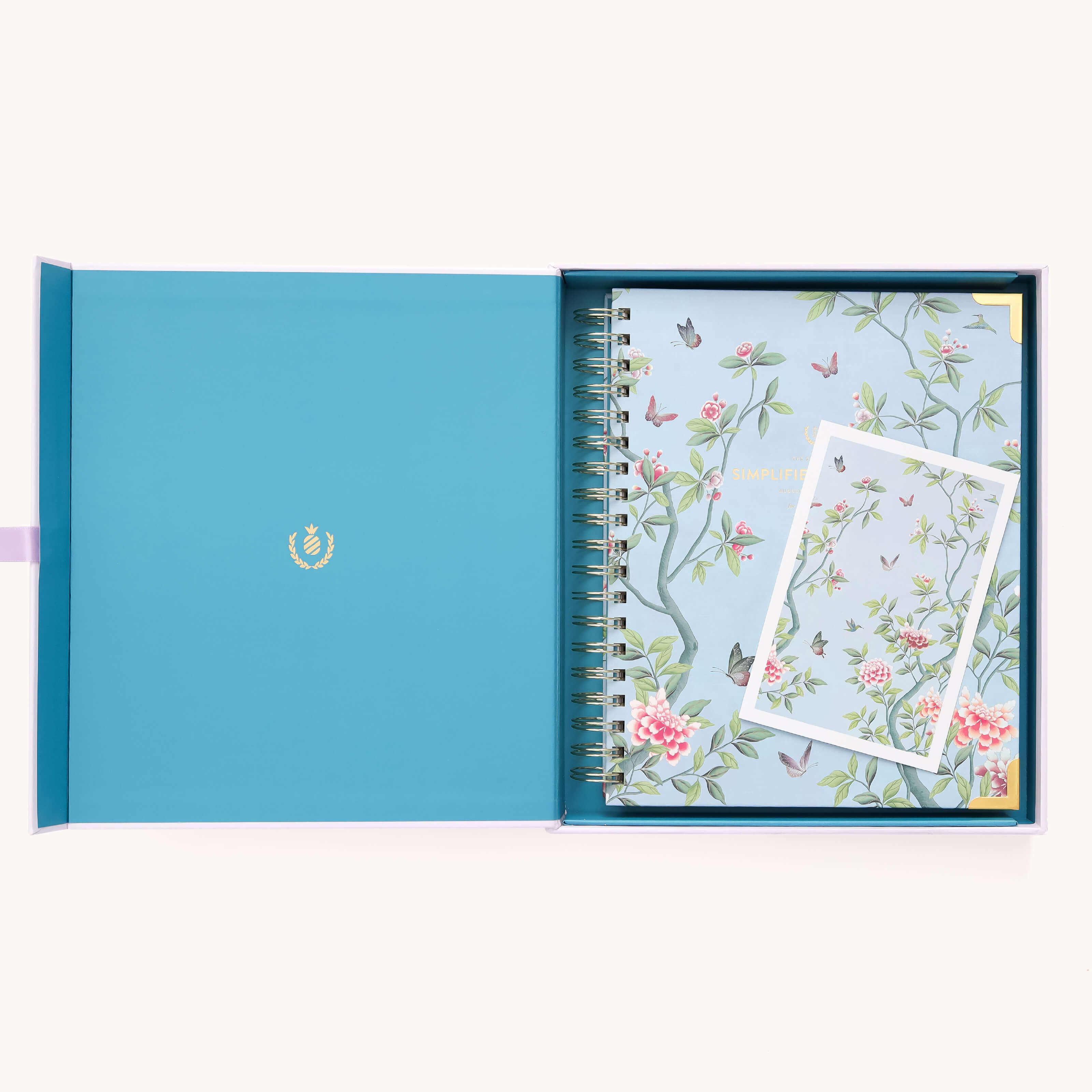 The 2025-2026 Spiral Weekly Simplified Planner - Faraway Land by Simplified features a floral-patterned notebook and matching card in a blue box. The spiral-bound notebook includes Prep Work pages, and the turquoise box is accented with an elegant gold foil emblem.