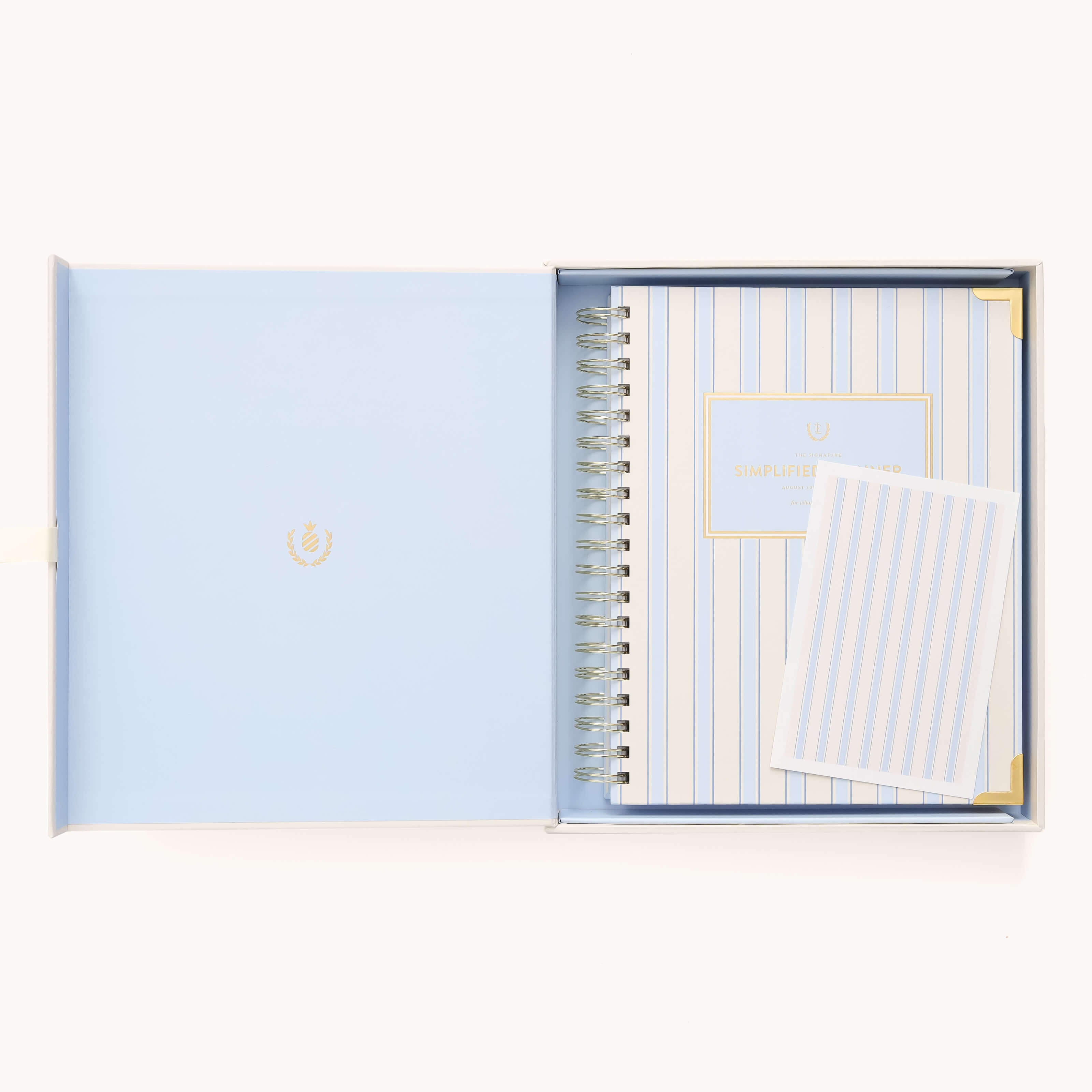 Open blue gift box with a 2025-2026 Spiral Weekly Simplified Planner - Serene Stripe featuring gold foil accents, plus a card. The planner boasts gleaming gold hues. The interior of the box is light blue with a decorative emblem, ideal for streamlined organization.