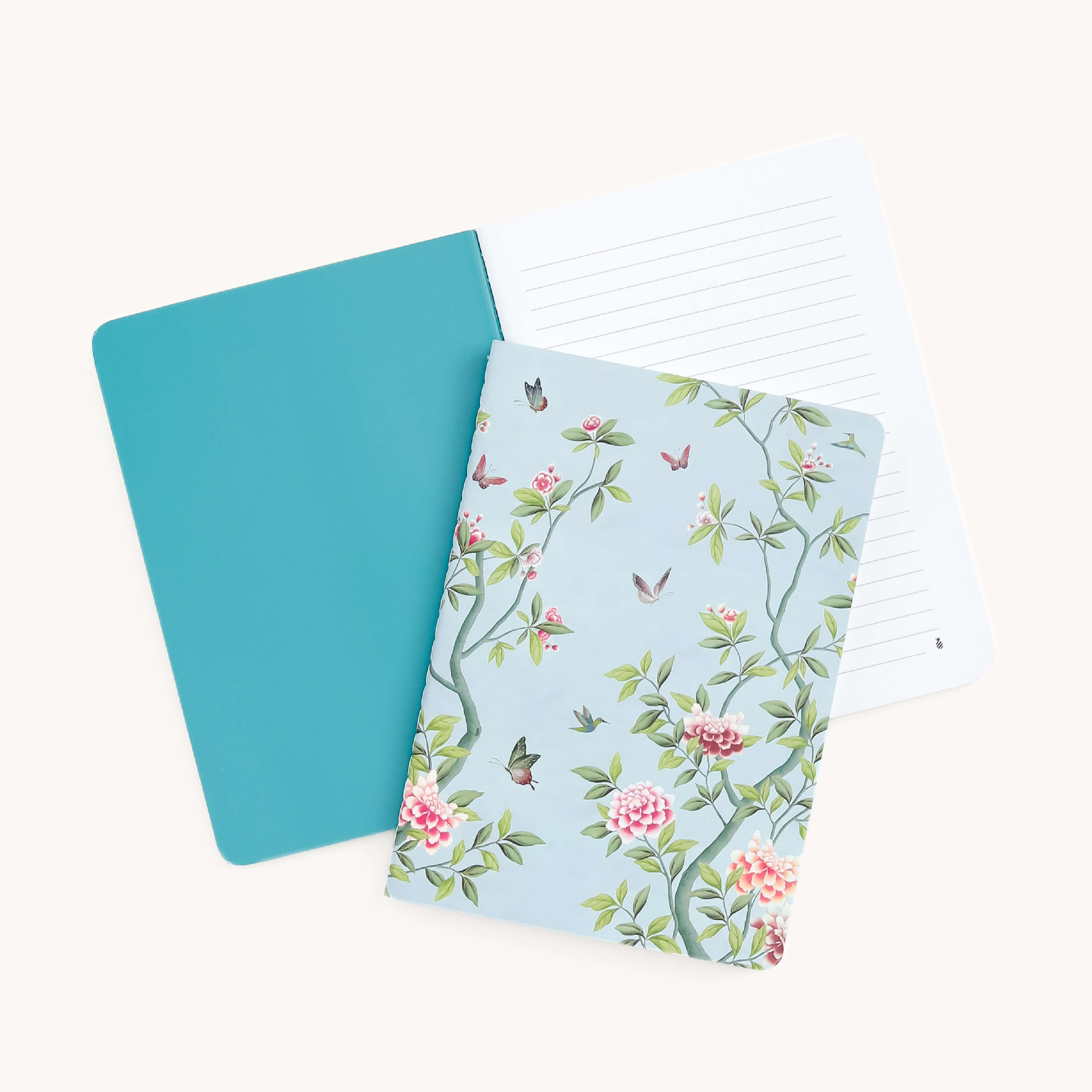 The two mini notebooks from Simplified include one with a teal cover and another with a custom floral and bird design on a blue background. Their thick paper covers and lined pages of the Mini Notebook - Faraway Land are visible as they’re displayed partially open.