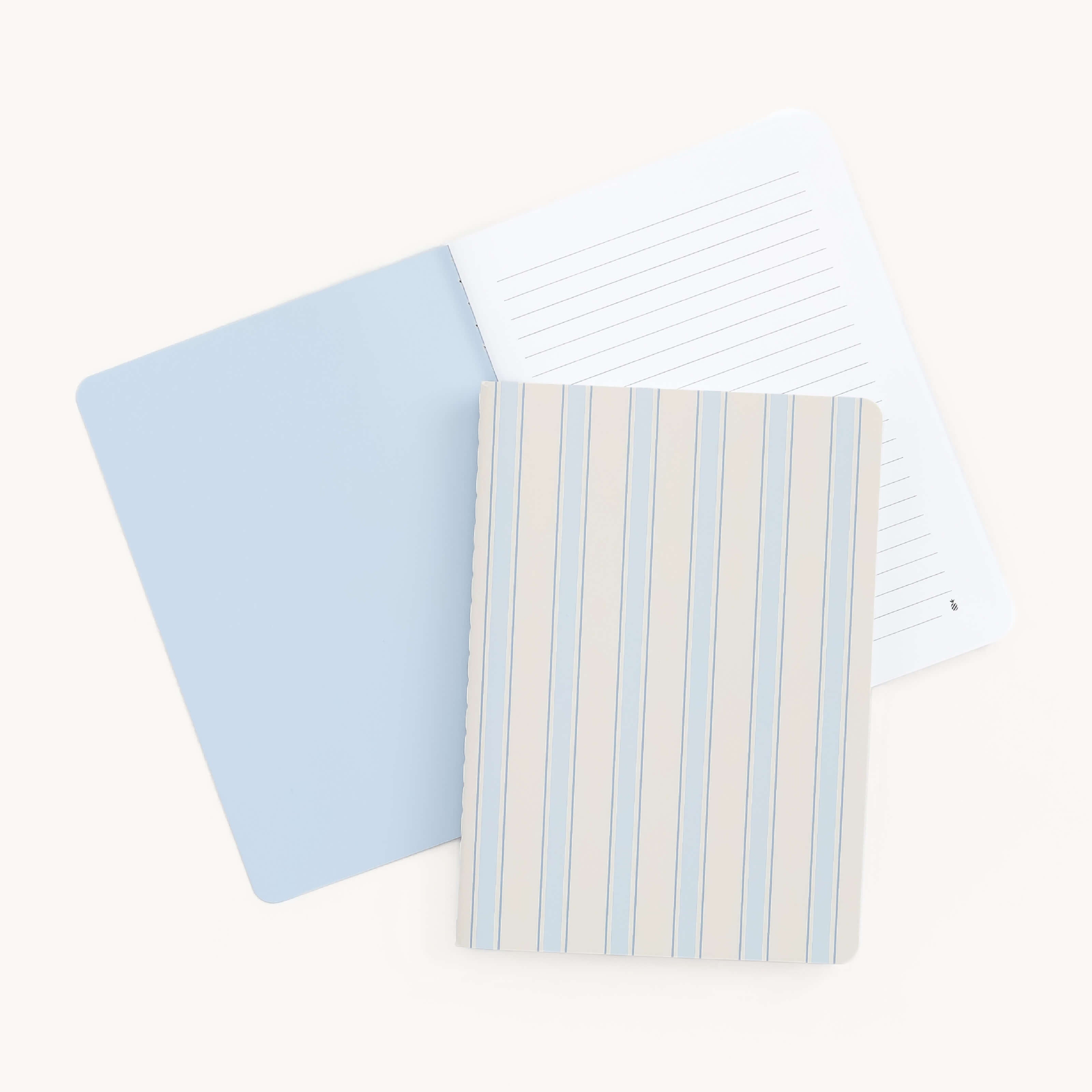 Two Mini Notebooks from Simplified rest on a white background. The top, named Serene Stripe, has a blue and beige striped cover, while the bottom is plain blue. The visible inner page of the bottom notebook is lined, ideal for notes on its thick paper cover.