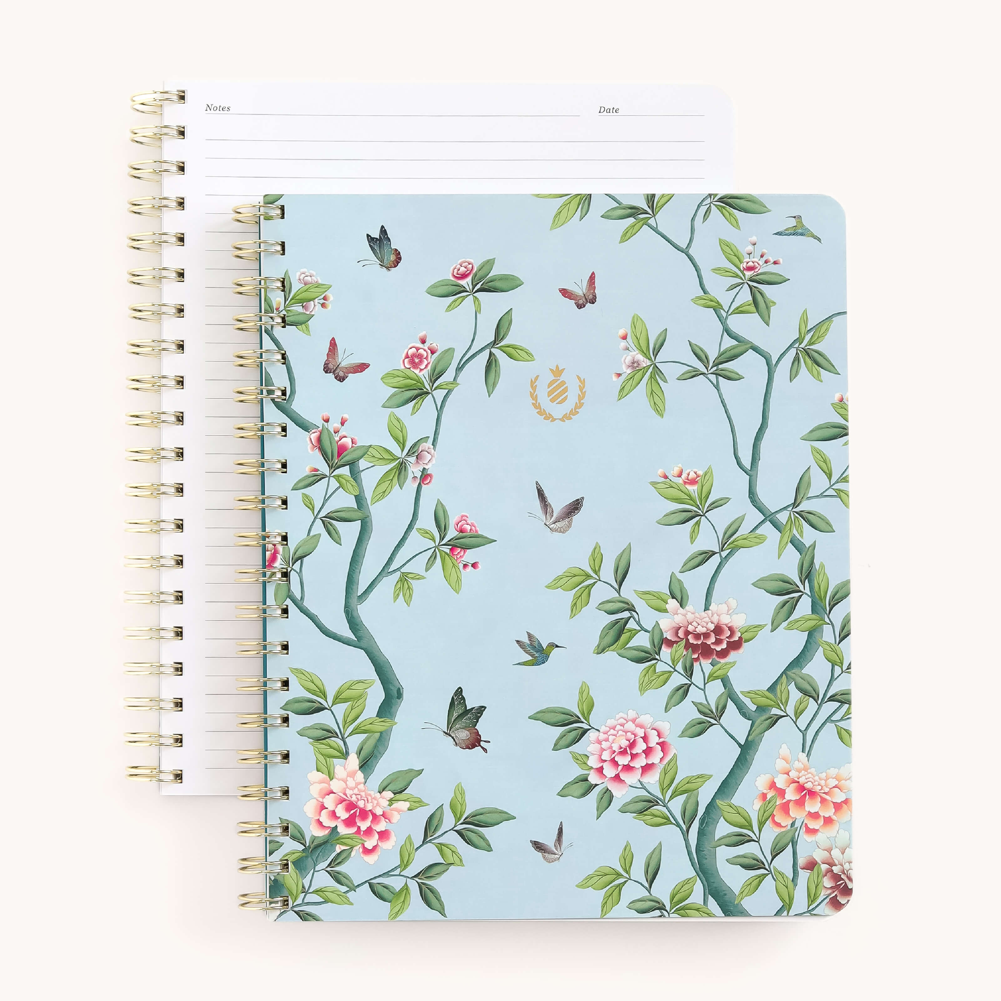 Two spiral-bound notebooks are stacked. The top, labeled Notebook - Faraway Land by Simplified, features a soft-touch cover with floral and hummingbird illustrations. Gold wire-o binding adds elegance, while the other notebook has lined pages with Notes and Date headers.