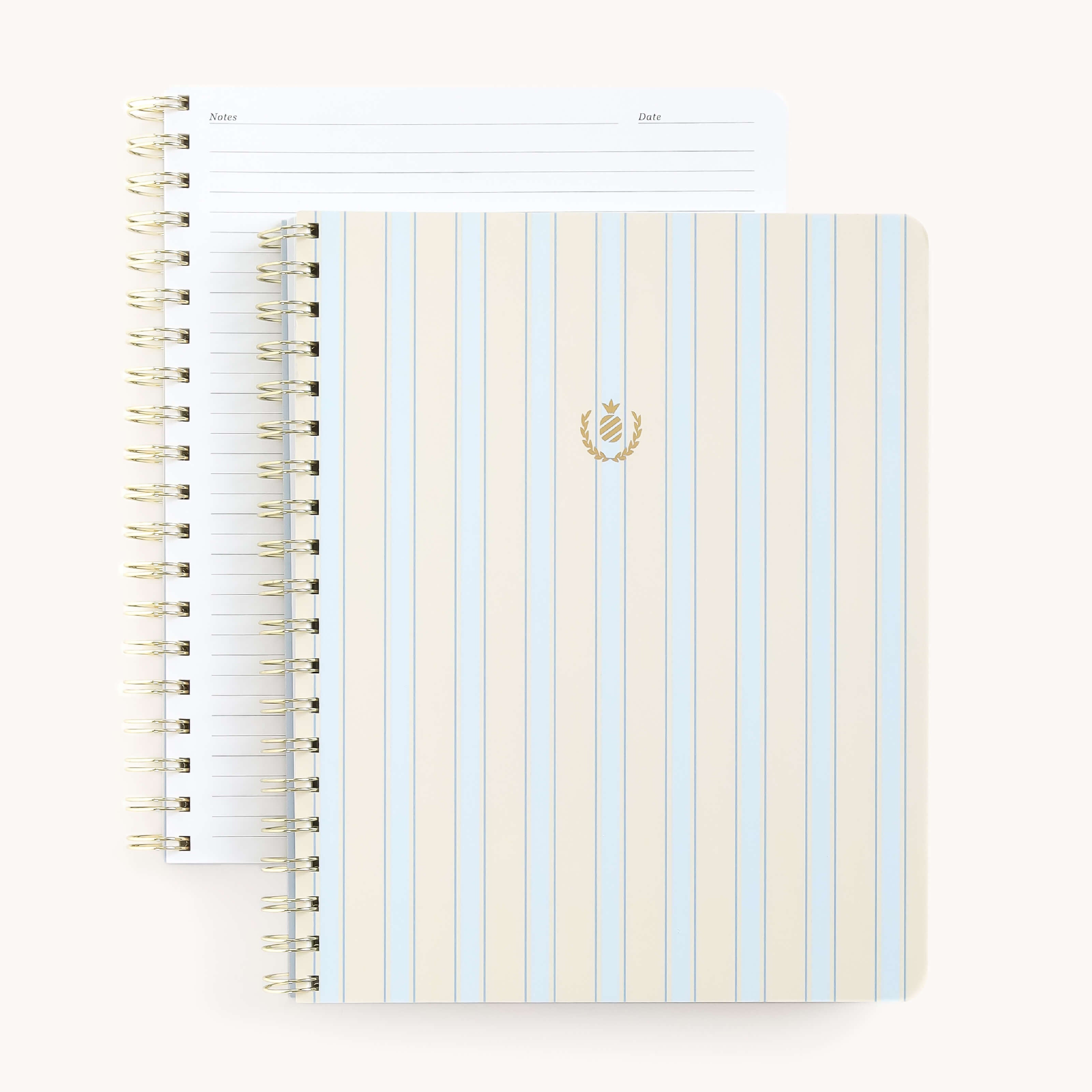 Two open spiral-bound notebooks rest on a white surface. The top one, Simplifieds Notebook - Serene Stripe, features a light striped soft-touch cover with a gold laurel emblem and sleek gold wire-o binding. The other notebook displays lined pages with headings for Notes and Date.