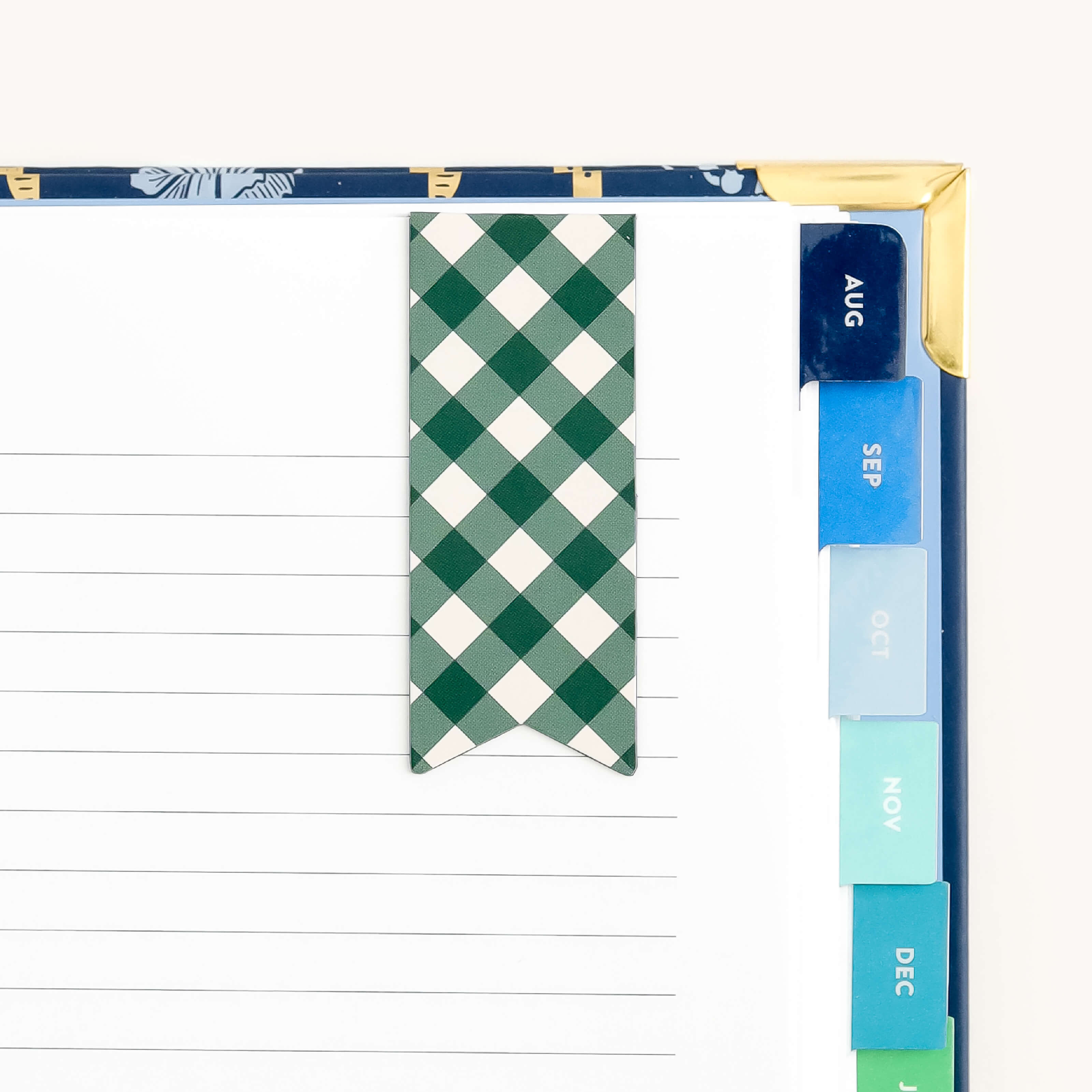 An open planner features a Simplified Page Marker in Camp Gingham design on a lined page. Month tabs from August to December appear as flag shapes in blue and green hues, while a gold corner adds elegance.