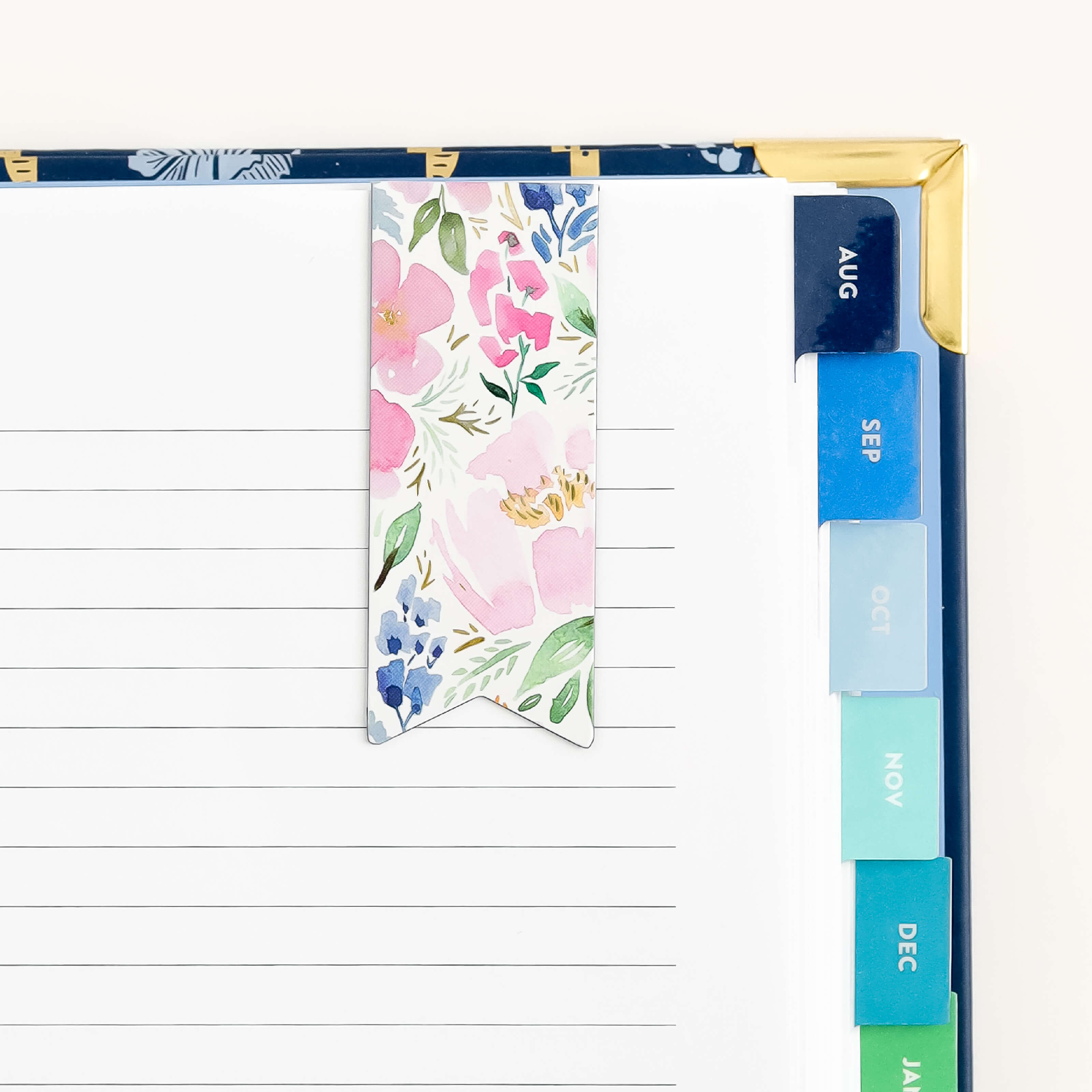 A close-up of a planner featuring the Cottage Floral magnetic bookmark by Simplified, opened to a lined page with blue and green tabs for August to December. The top right corner is adorned with a gold metal protector.
