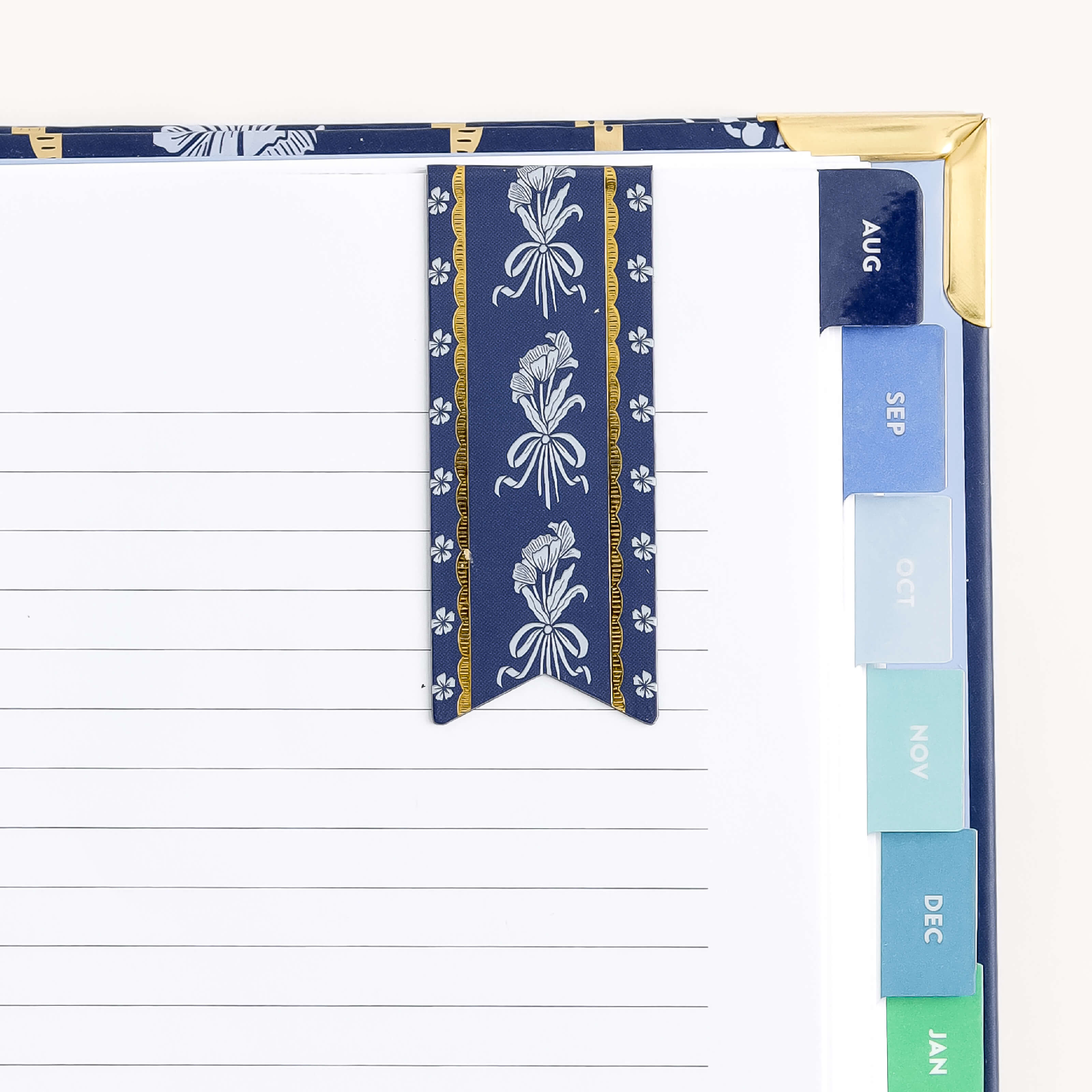 The Simplified Poppy Posies planner, with lined pages and monthly tabs from August to January, features a floral-patterned blue ribbon bookmark. Its cover corners are reinforced with gold and it includes a flag-shaped magnetic page marker for added convenience.