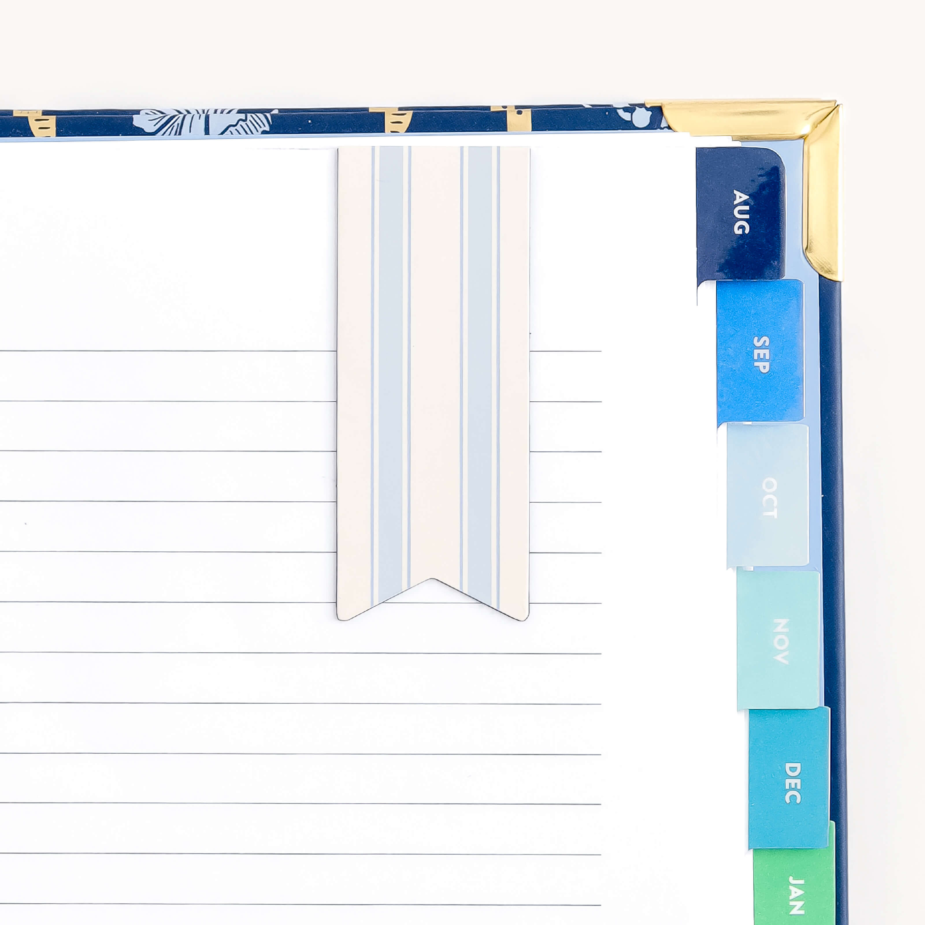 This Simplified planner features lined pages, a vibrant Serene Stripe bookmark, and monthly tabs from August to January in gradient blues and greens. The cover is elegantly detailed in gold, while a magnetic page marker enhances its convenience and style.