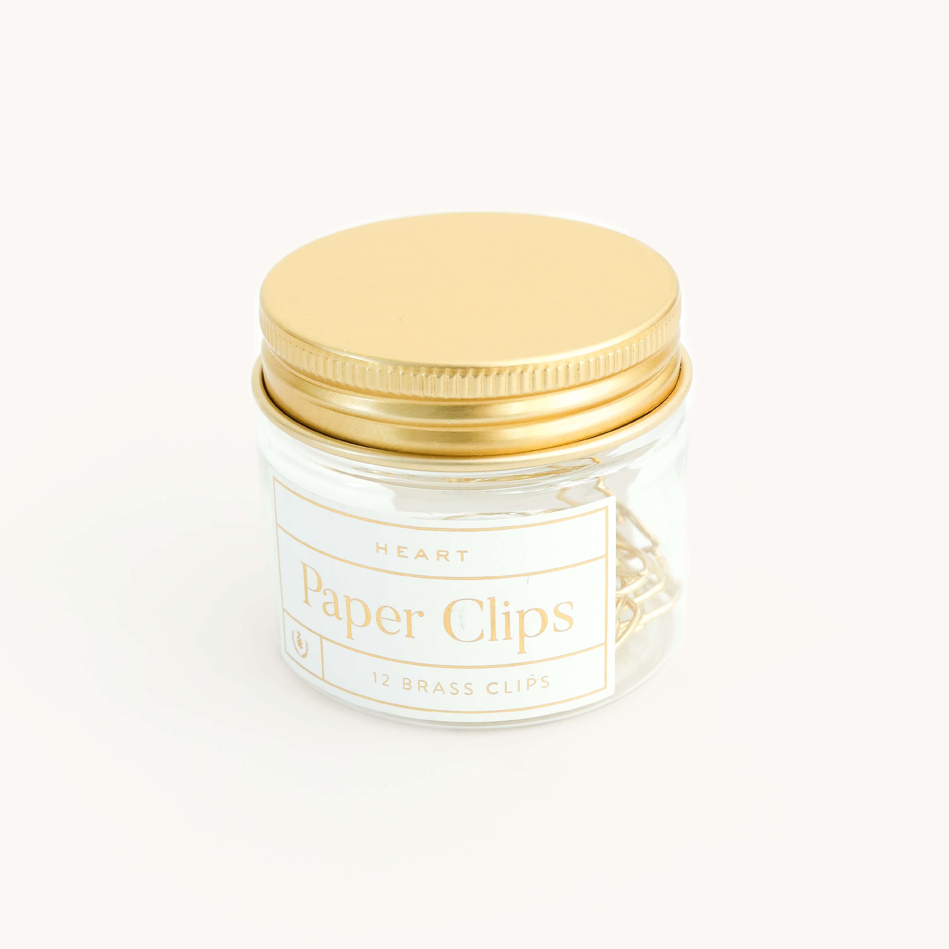 A clear glass jar with a gold screw-on lid contains durable metal heart-shaped paper clips. The label reads “Paper Clips - Heart, Simplified® by Emily Ley, 12 Brass Clips.”.