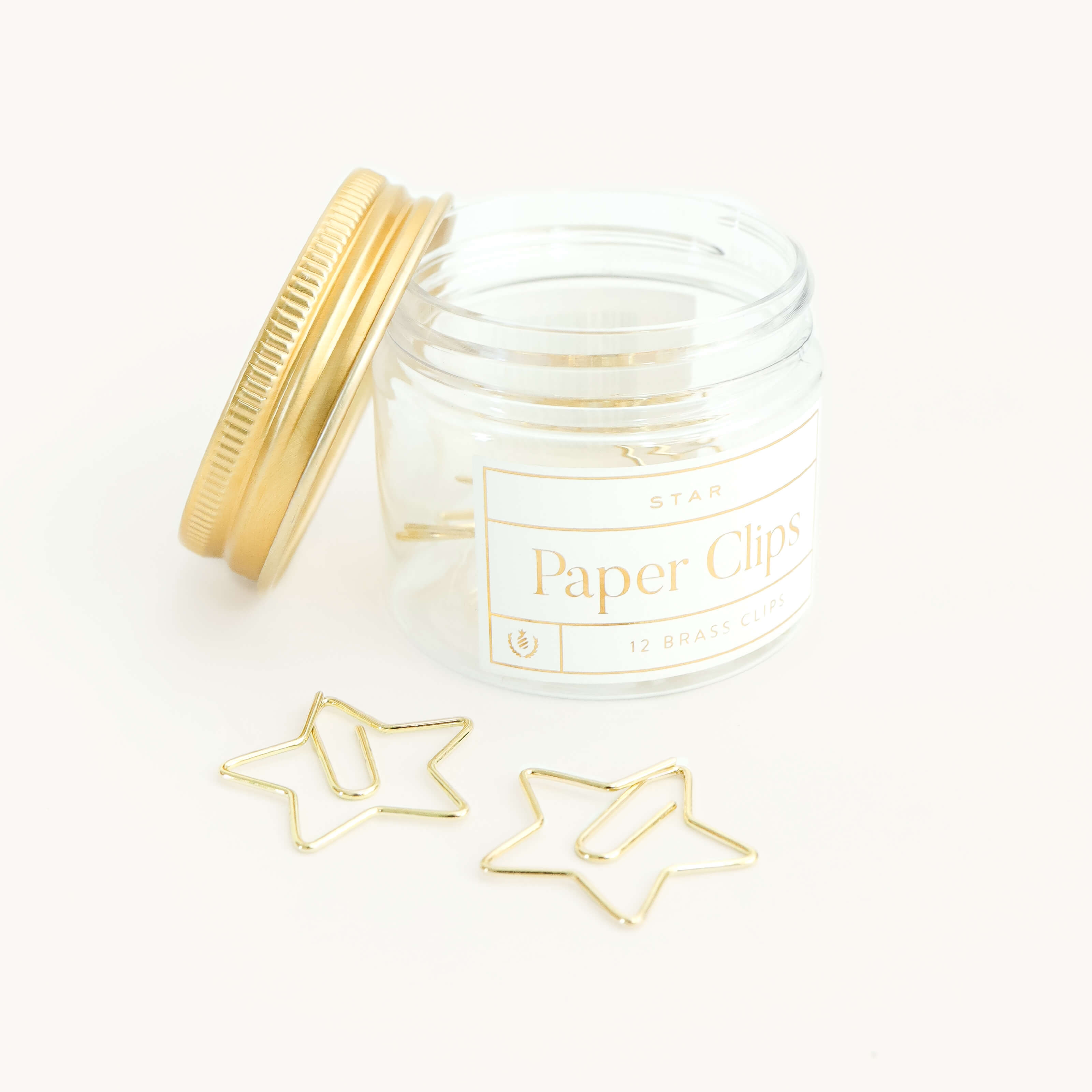 An open jar labeled "Simplified® by Emily Ley Paper Clips - Star, 12 Brass Clips" displays two star-shaped clips on a white surface, bringing playful elegance to your workspace.