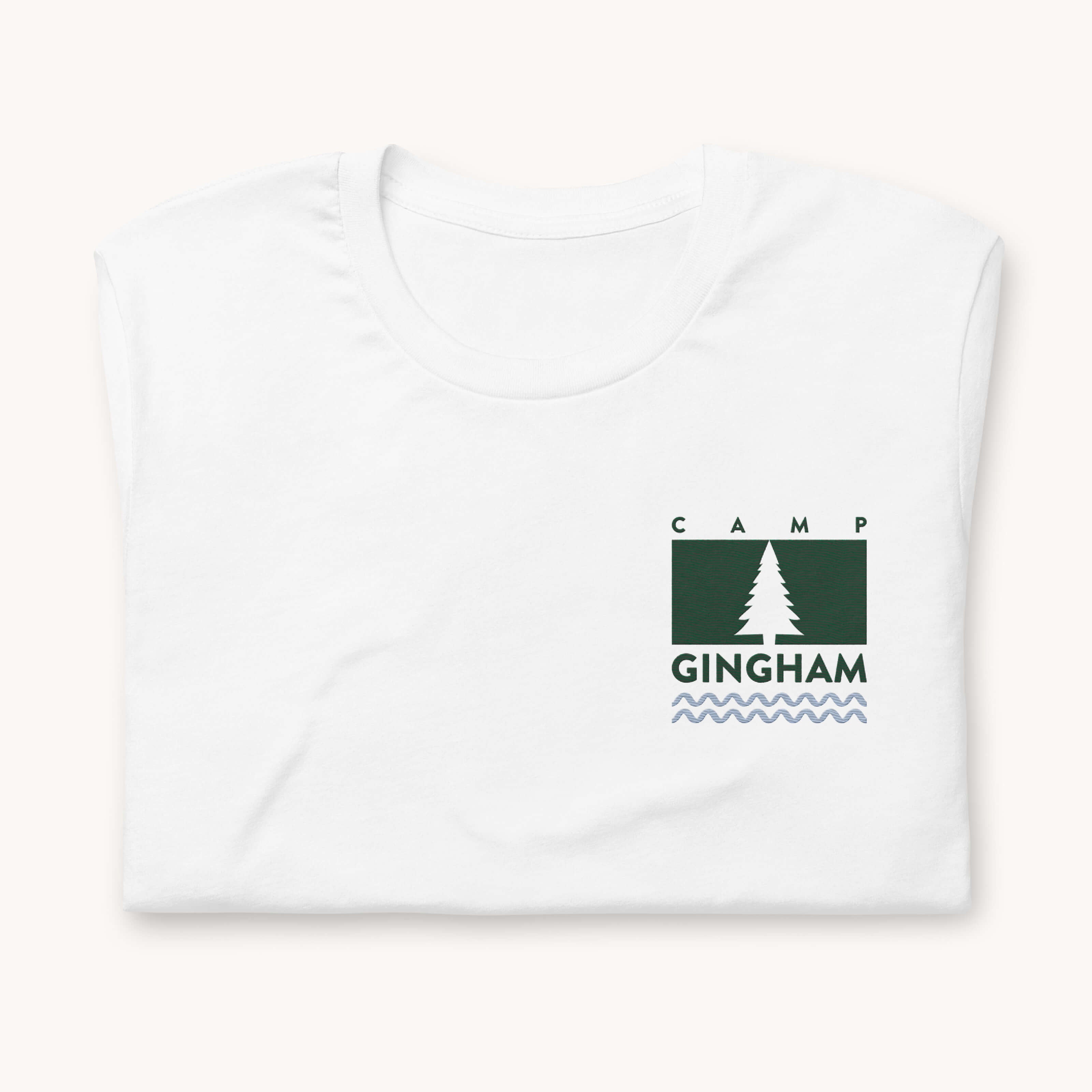 The Simplified Embroidered T-Shirt - Camp Gingham features a folded white fabric with a green rectangular logo, white pine tree, CAMP GINGHAM text, and a wavy blue line embroidered on the upper left chest for classic camp style.