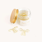 A small glass jar with a gold lid holds Simplifieds Paper Clips - Bows, featuring durable, bow-shaped metal brass clips. Two clips rest on the white surface outside the jar. The label reads Bow Paper Clips, 12 durable metal brass clips.