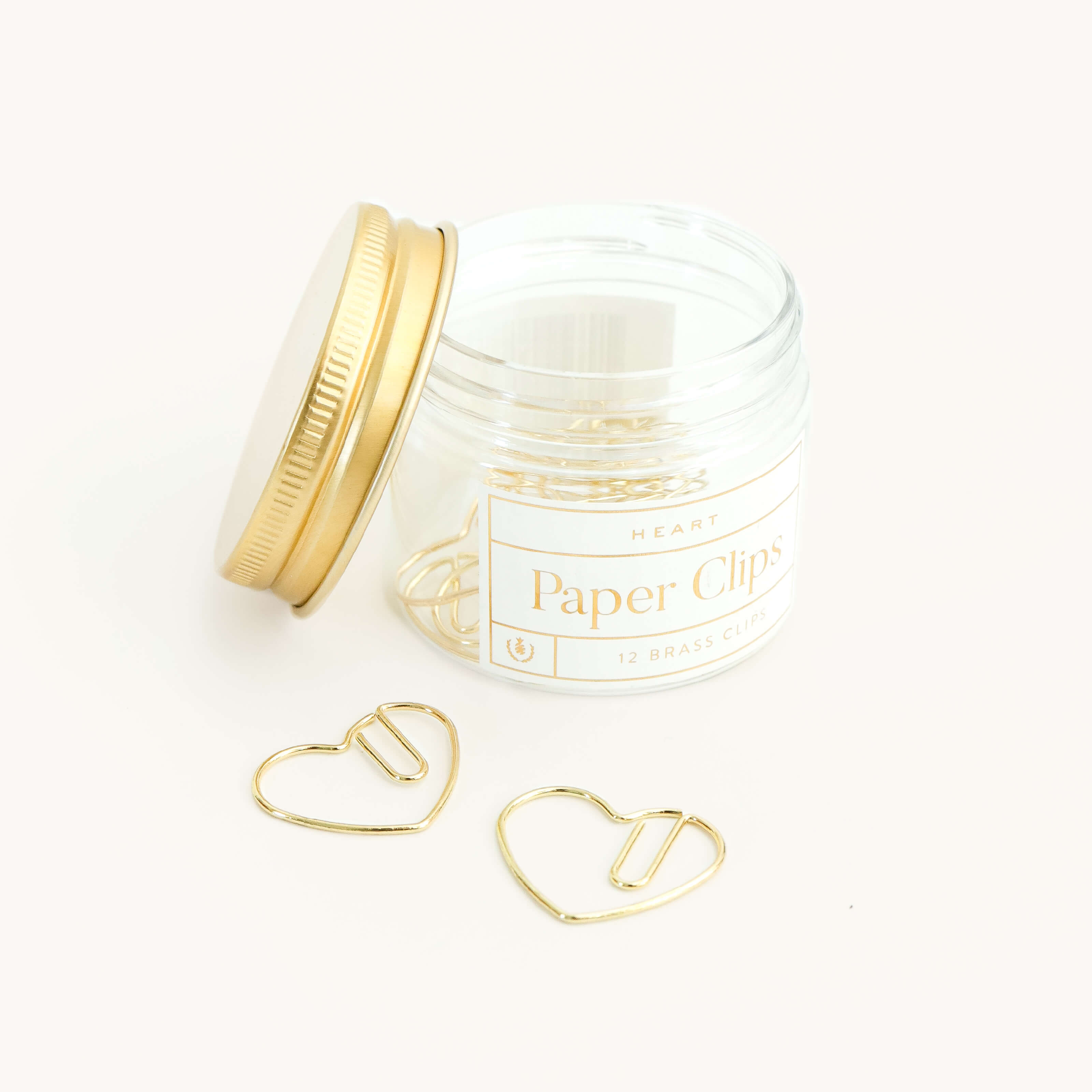 A clear jar labeled "Paper Clips - Heart" by Simplified® by Emily Ley, topped with a gold lid and filled with durable, heart-shaped metal paper clips. Two heart paper clips rest outside the jar on a white surface.