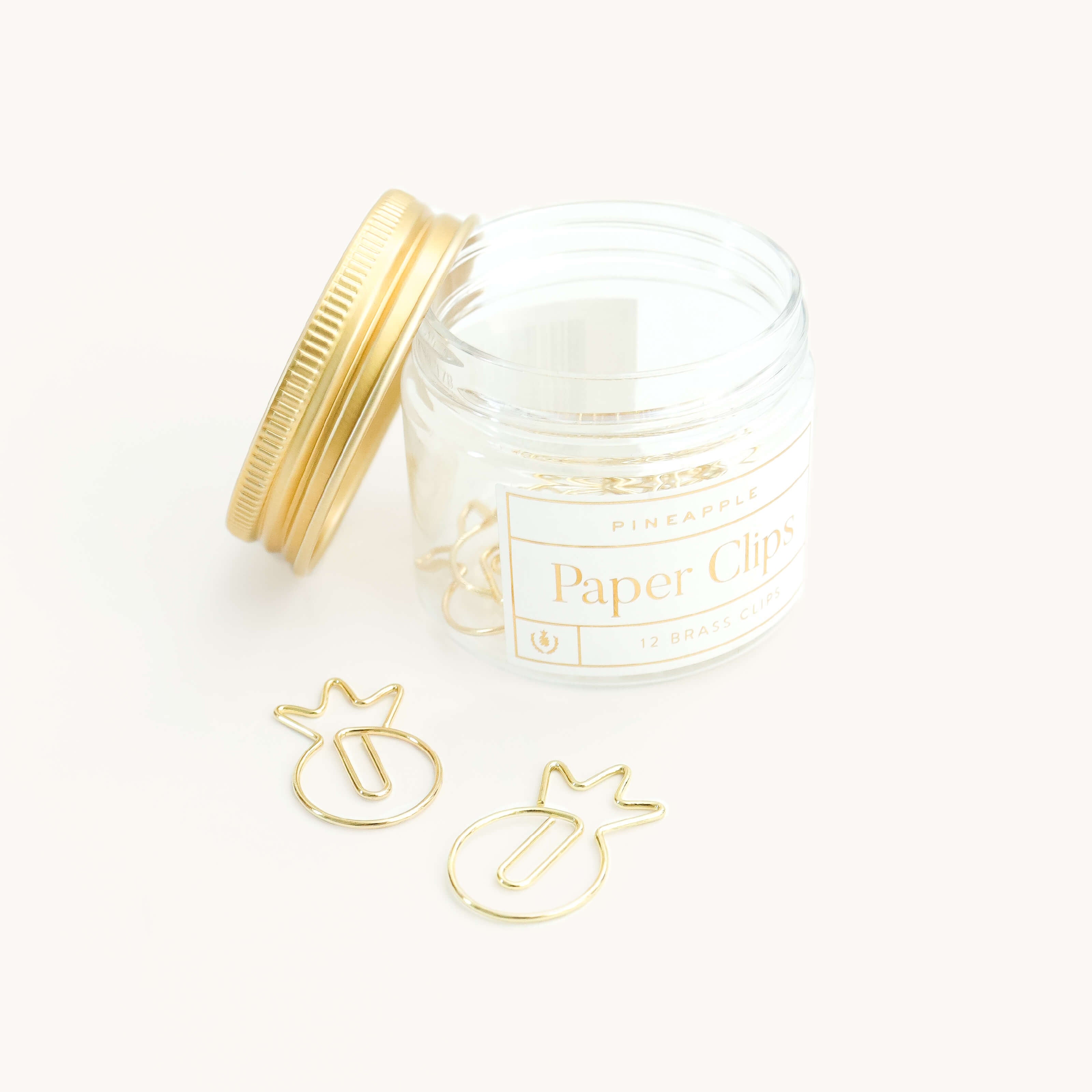 A clear jar labeled "Paper Clips - Pineapples" by Simplified sits with its gold lid nearby, holding gold pineapple-shaped paper clips. Two shaped clips are displayed outside the jar on a white surface.