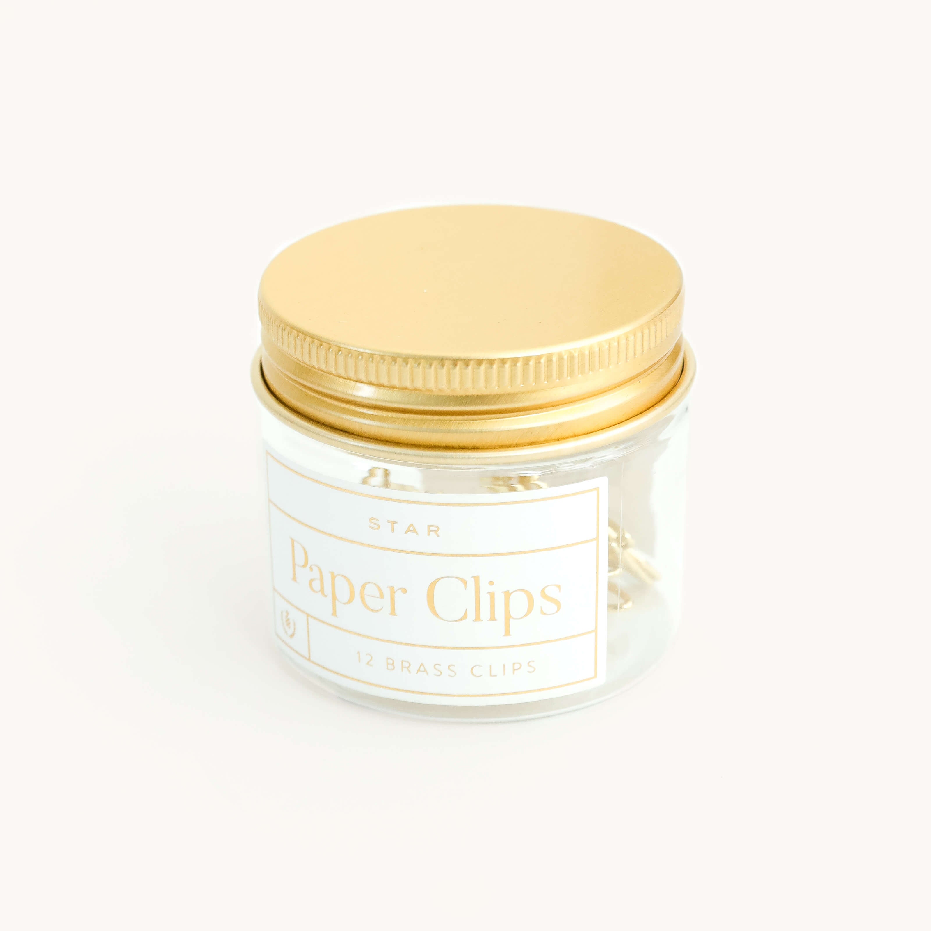 A clear glass jar with a gold lid holds 12 brass star-shaped paper clips. The label reads "Paper Clips - Star" by Simplified® by Emily Ley, combining elegance and function for anyone seeking stylish organization.