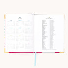 The 2025-2026 Bookbound Weekly Simplified Planner by Simplified opens to reveal a calendar on the left and a holiday list on the right. A light blue ribbon bookmark is centered, with subtle gold foil edging. The plain white background enhances its minimalist elegance.