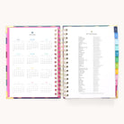 The 2025-2026 Spiral Daily Simplified Planner by Simplified is open showing a calendar on the left and holidays on the right. It features colorful tabbed dividers, gold foil details, is spiral-bound, and displayed against a plain background.