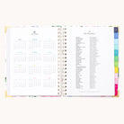 The Simplified 2025-2026 Spiral Weekly Planner in Cottage Floral presents a left-side calendar and right-side holiday list. It boasts vibrant tabbed sections, floral borders, and gold foil details for an elegant touch.