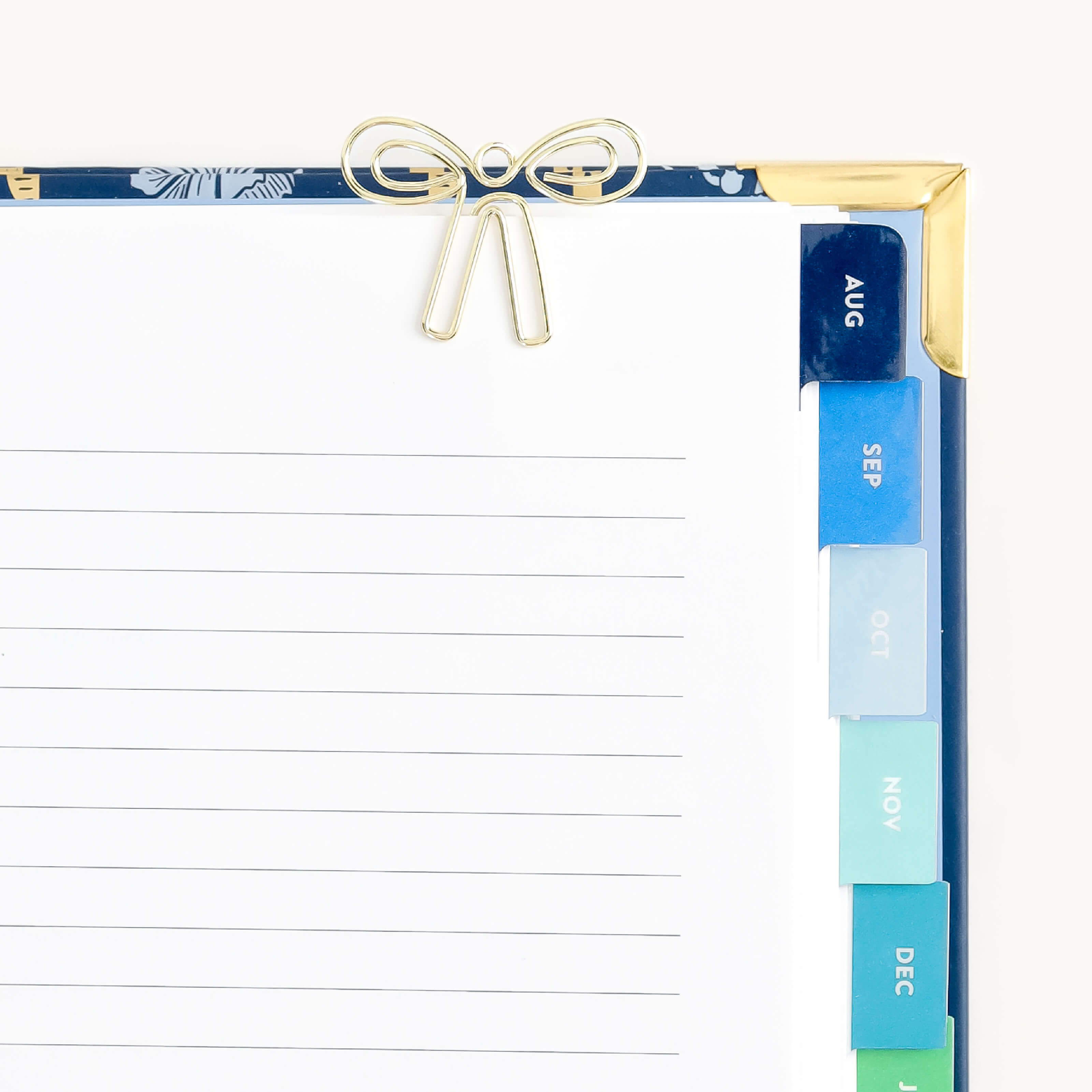 A decorative clipboard by Simplified features a durable metal bow-shaped paper clip holding lined paper. Colorful monthly tabs from August to January line the right edge. It boasts a floral design with gold corners. The product used for securing papers is called Paper Clips - Bows.