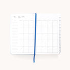 The Simplified 2025-2026 Bookbound Daily Dapperdesk Planner in Carolina Dot, featuring a leatherette cover, showcases an August 2025 monthly calendar. It includes a blue ribbon bookmark with dates from Sunday to Saturday and ample note space on the left, against a plain white background.