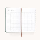 The Simplified 2025-2026 Bookbound Daily Dapperdesk Planner in Evergreen offers a Sunday-start August 2025 monthly calendar, with a brown ribbon bookmark. Opposite the grid are lined pages for detailed notes, and tabs on the right edge help navigate each month smoothly.
