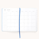 The Simplified 2025-2026 Bookbound Weekly Dapperdesk Planner in Carolina Dot features a monthly calendar starting August 2025, notes on the left and weekly days on the right. It includes a blue ribbon bookmark, right-edge tabs, and a sleek leatherette cover.