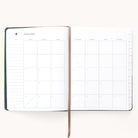 The Simplified 2025-2026 Bookbound Weekly Dapperdesk Planner - Evergreen elegantly showcases an August 2025 monthly calendar. The left page offers space for notes, the right features a Sunday-to-Saturday grid, and a brown ribbon bookmark marks the center of this refined 12-month planner.