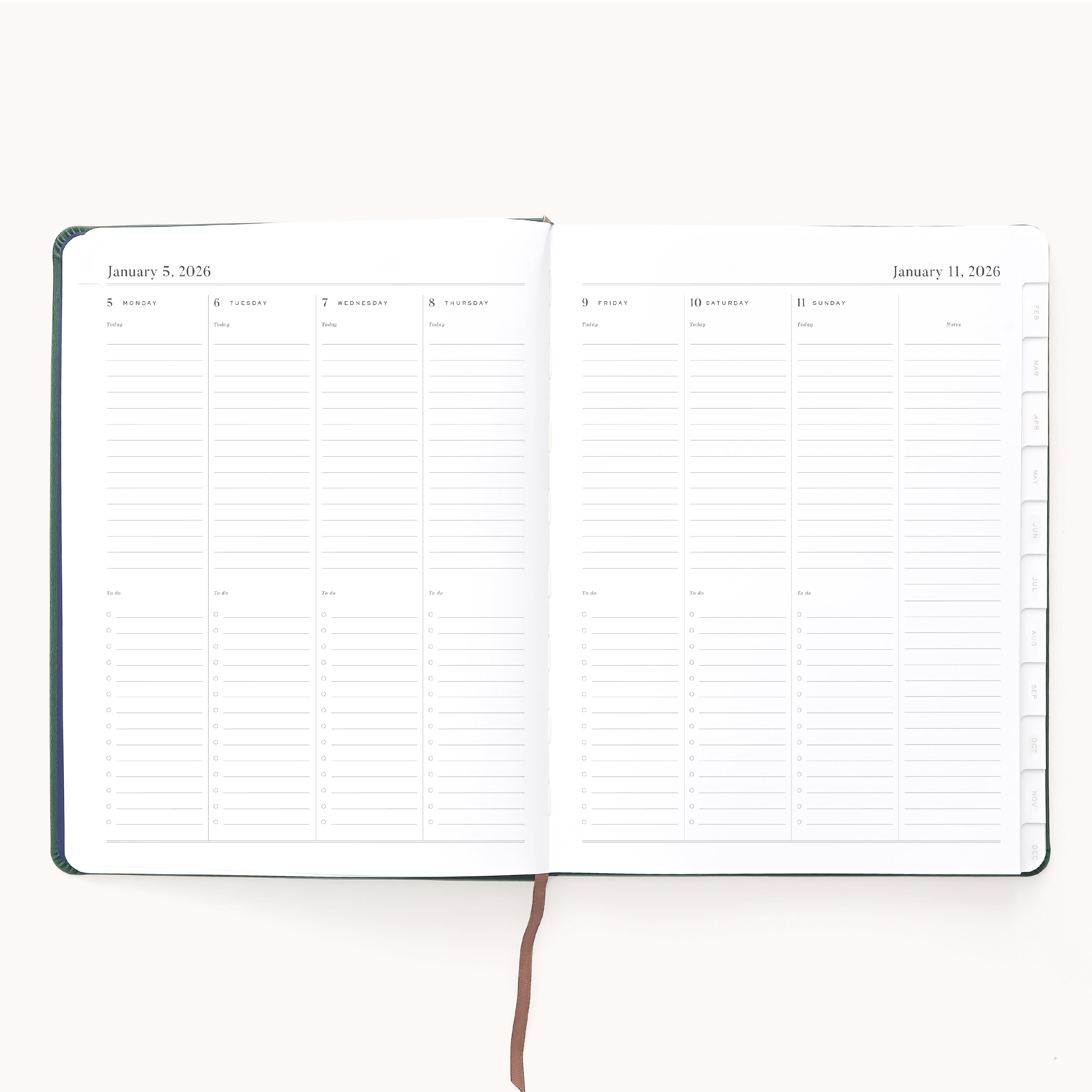 The Simplified 2026 Bookbound Weekly Dapperdesk Planner in Evergreen lies open, showing January 5–11 with daily columns, lined note sections, and a brown ribbon bookmark resting on its elegant leatherette cover.