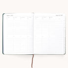 The Simplified 2025-2026 Bookbound Weekly Dapperdesk Planner in Evergreen, with a leatherette cover, features a layout from August 4 to August 10, 2025. Each days ruled sections accommodate time slots and notes, marked by a central brown ribbon bookmark.