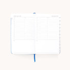 The 2025-2026 Bookbound Daily Dapperdesk Planner - Carolina Dot by Simplified is open, revealing a leatherette cover and a spread for Thursday, August 3, and Friday, August 4. The pages feature an hourly schedule from 7 AM to 8 PM plus a notes section and a centered blue ribbon bookmark.
