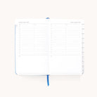 The Simplified 2025-2026 Bookbound Daily Dapperdesk Planner in Carolina Dot features a daily layout, including hourly slots from 7 AM to 7 PM, notes section, and a blue ribbon bookmark. Its leatherette cover opens to reveal an organized schedule for August 19-20, 2023.