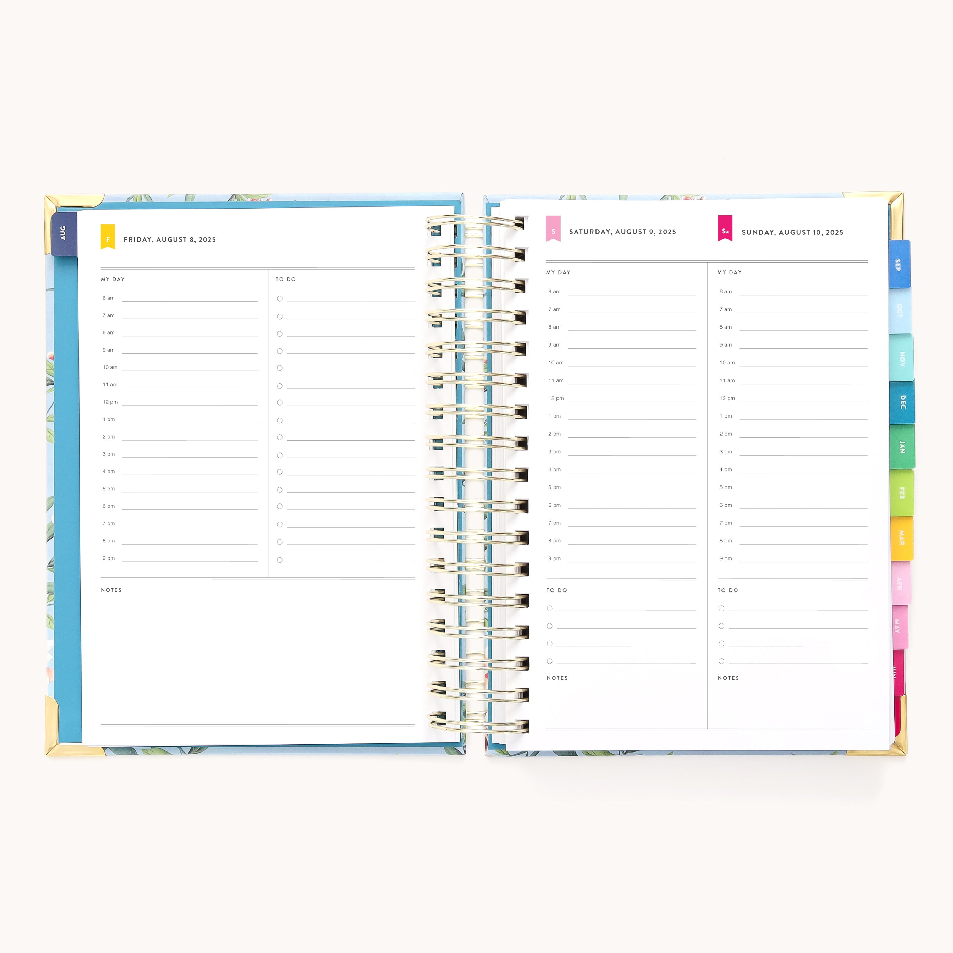 The 2025-2026 Spiral Daily Simplified Planner in the Faraway Land design is open, showing pages for August 7-9, 2023. Each page features a lined schedule with time slots and colorful tabbed dividers. Its cover boasts a blue floral pattern enhanced with elegant gold foil details.