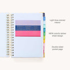 The 2025-2026 Spiral Daily Simplified Planner in Serene Stripe by Simplified has light blue blank pages, colorful labeled tabs on the right, vibrant sticky notes with labels and icons on the left, and features gold corner protectors with elegant gold foil details.