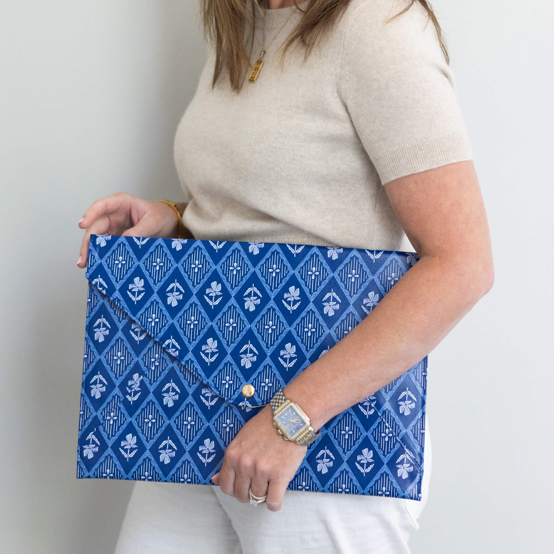 A woman in a beige short-sleeve top and white pants holds the Simplified Leatherette Tech Case - Block Floral, featuring a blue patterned design and magnetic closure. She accessorizes with a gold necklace and silver watch against a plain background.