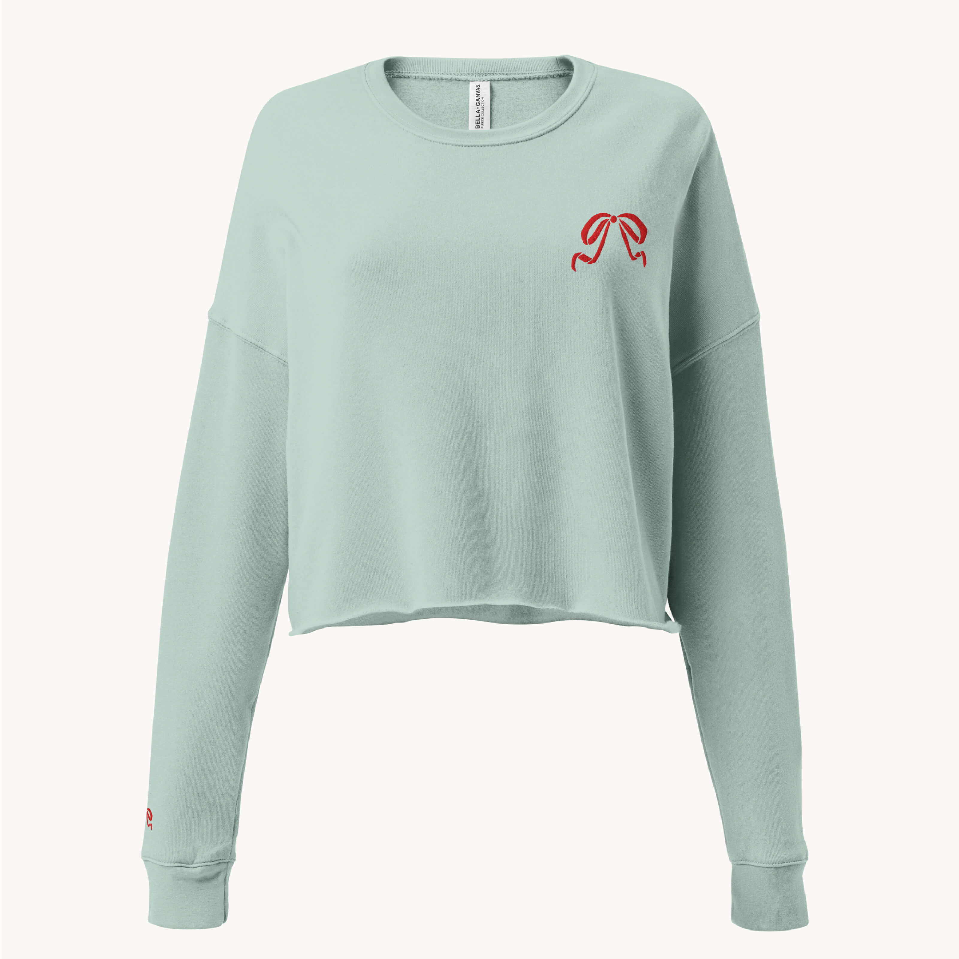 The Simplified Embroidered Crop Sweatshirt - Red Bow is a light green, long-sleeve cropped top featuring a small red bow embroidered on the upper left chest and a tiny red heart near the left sleeve cuff.